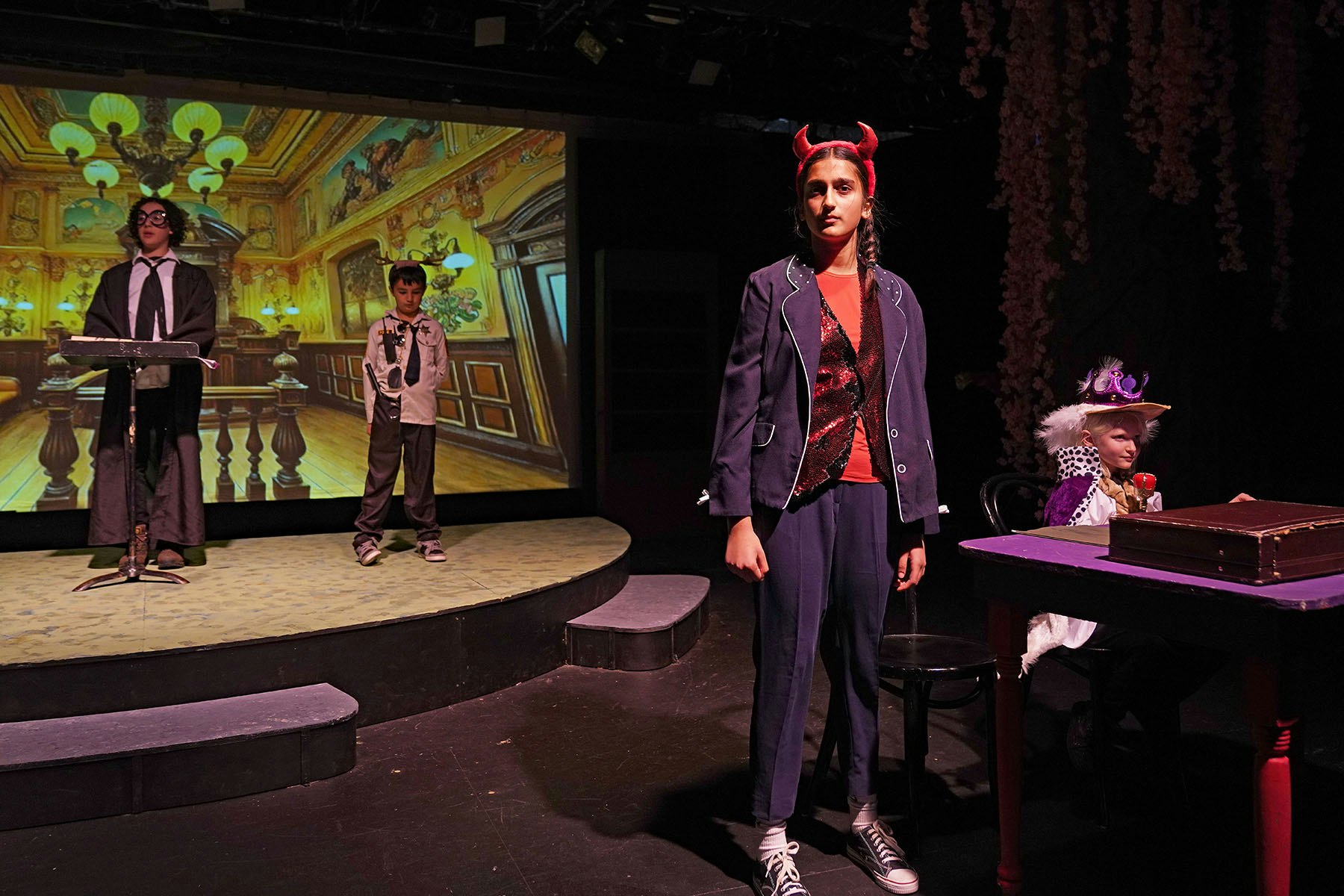 A stage performance with four children in costumes; one wears devil horns, and another a crown, in front of a projected backdrop.