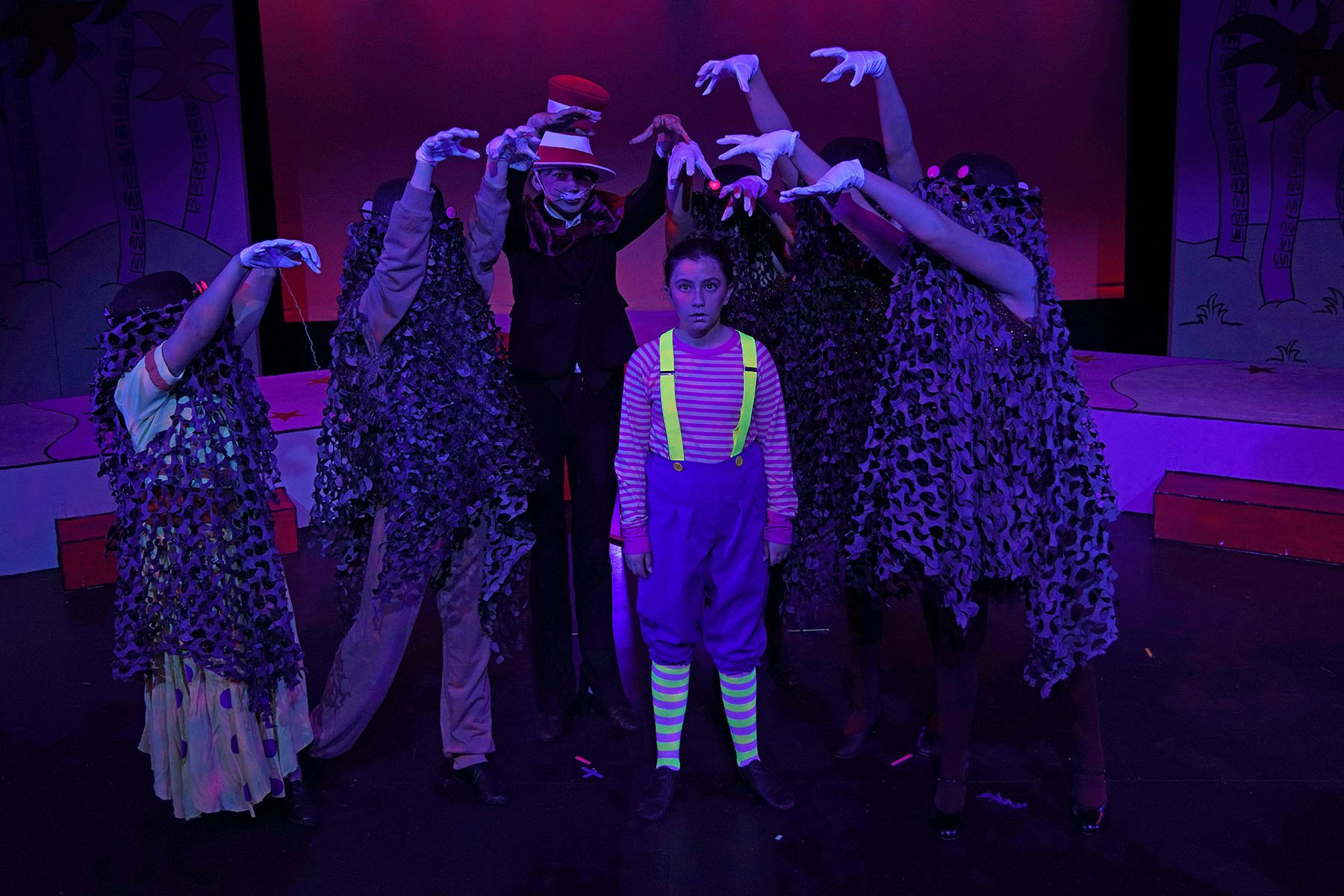 A theatrical scene featuring a central child surrounded by characters in dark costumes, with vibrant lighting and dramatic poses.