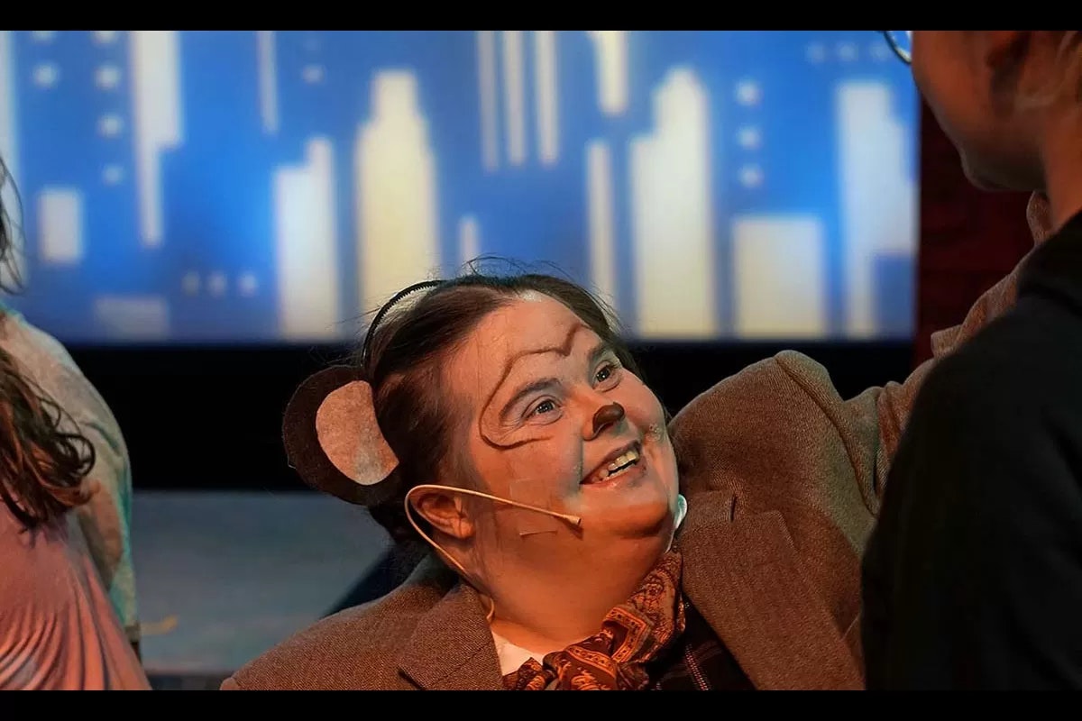 A person in animal makeup and costume, likely performing on stage with a cityscape backdrop, smiling at someone.