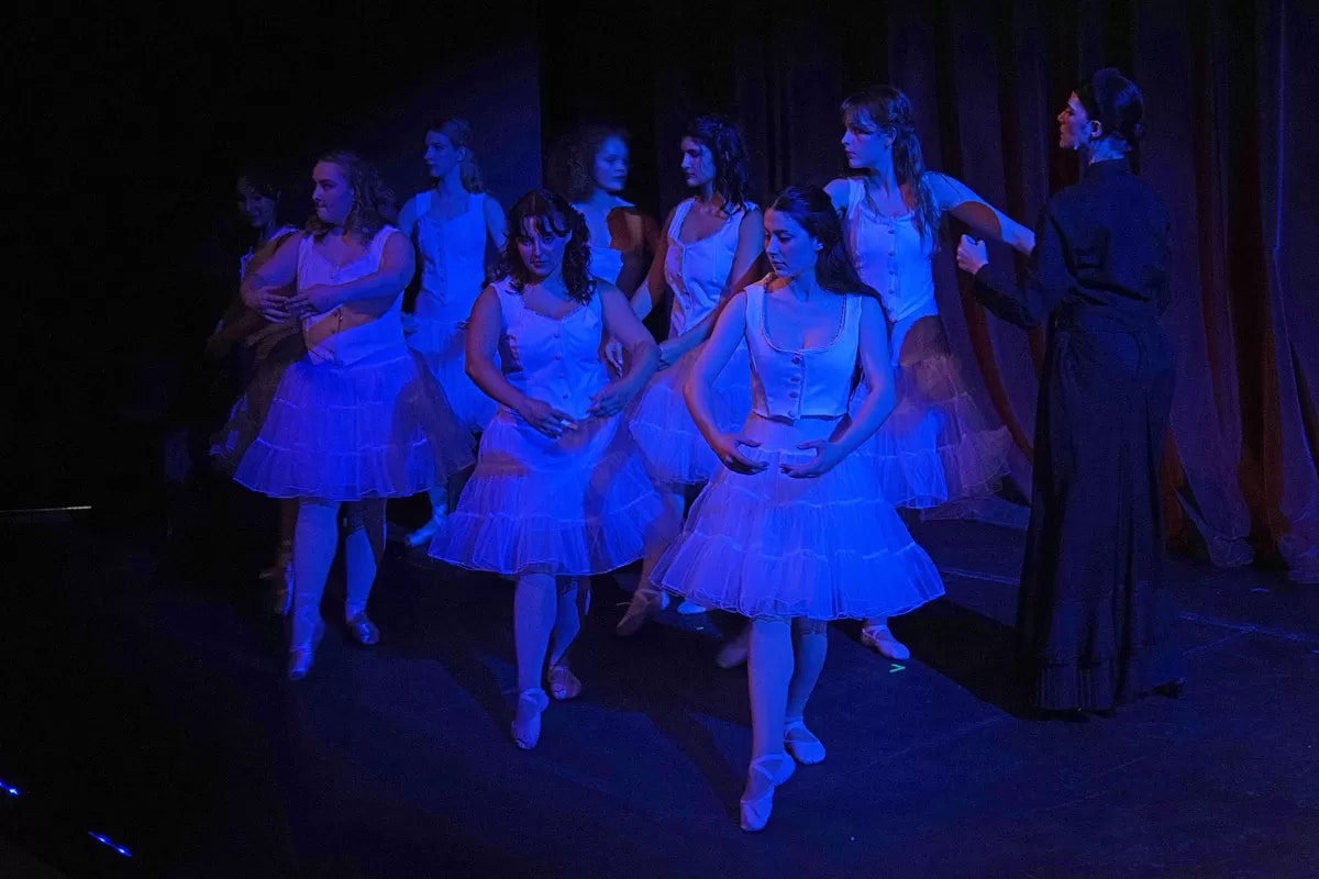Ballerinas in white tutus rehearsing or performing on stage under blue lighting.