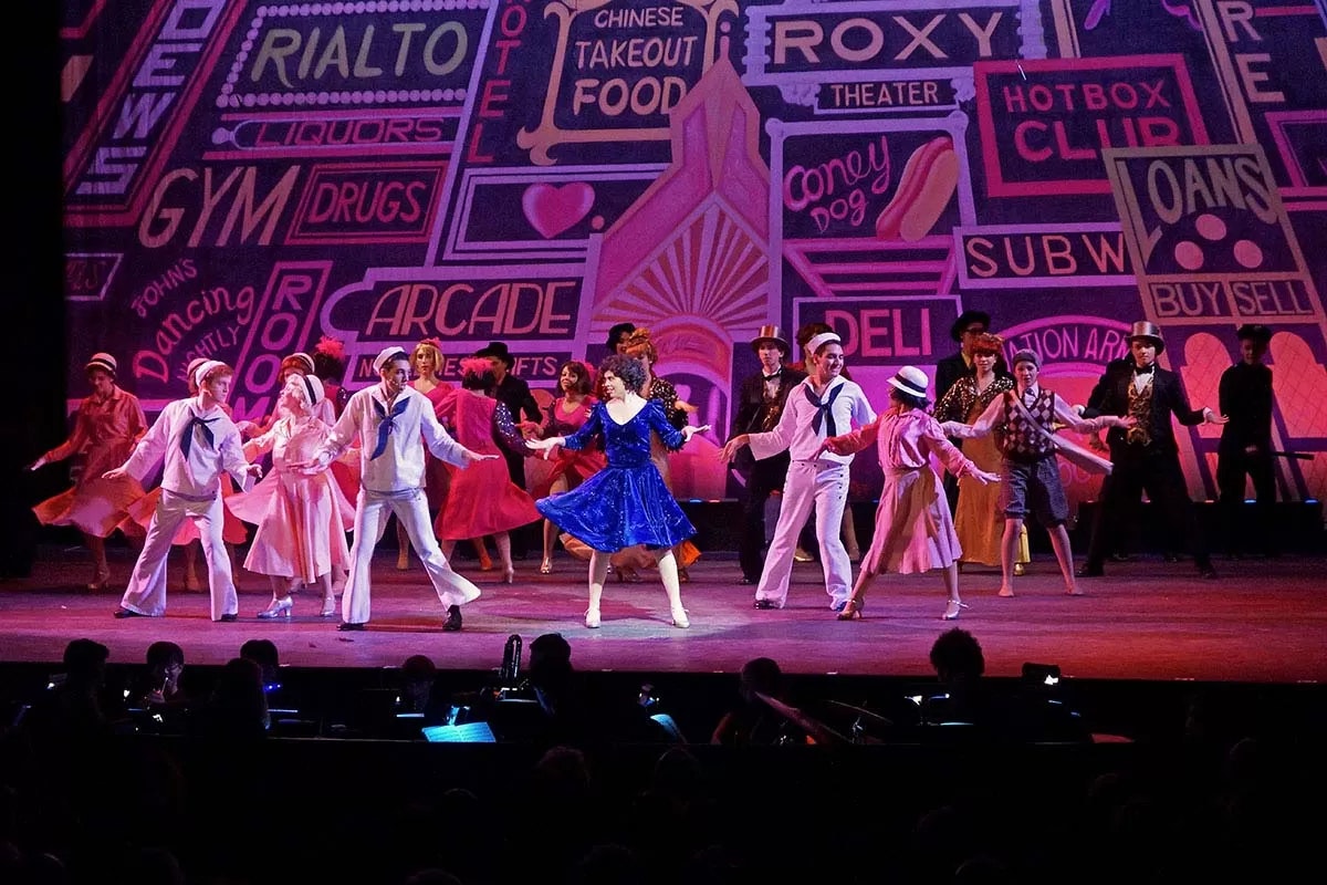 A lively stage performance with dancers in various costumes, colorful neon sign backdrop displaying words like "Rialto," "Arcade," and "Deli."