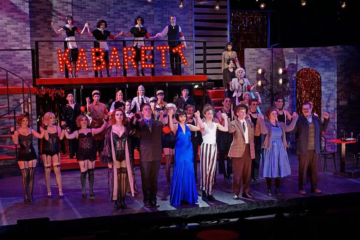 A group of performers takes a bow on stage in front of a "KABARETT" sign, dressed in theatrical costumes.