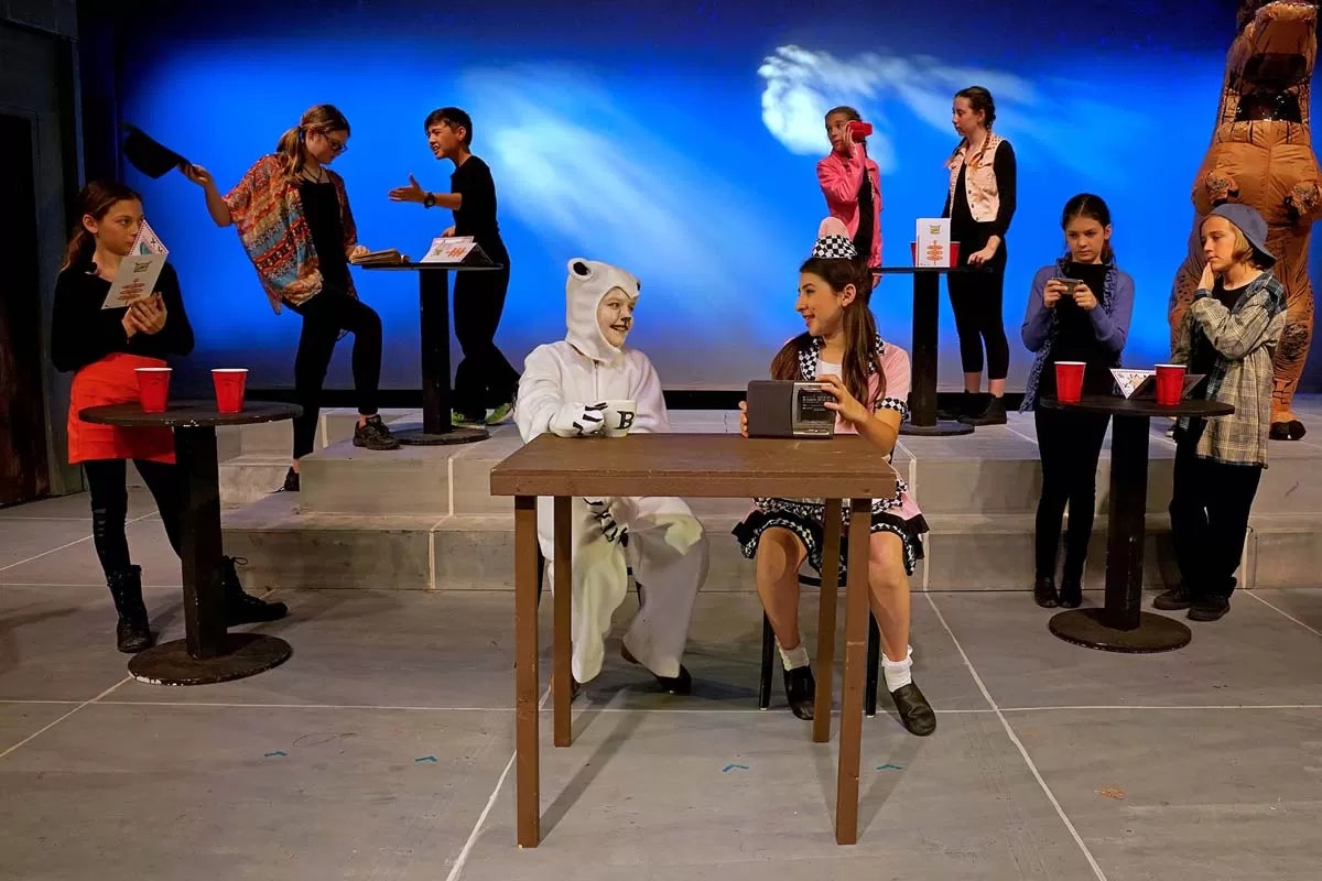 Children performing on stage in costumes, engaging in a play scene with tables and cups against a blue backdrop.