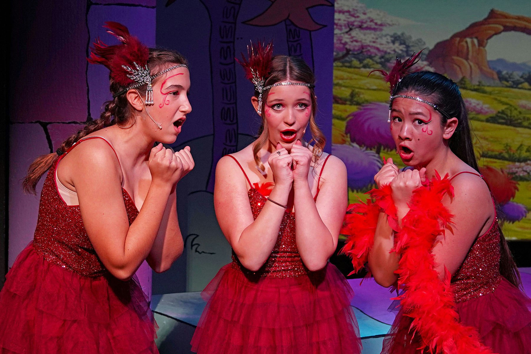 Three young performers in sparkling red dresses express surprise or excitement during a stage performance, with dramatic makeup and accessories.