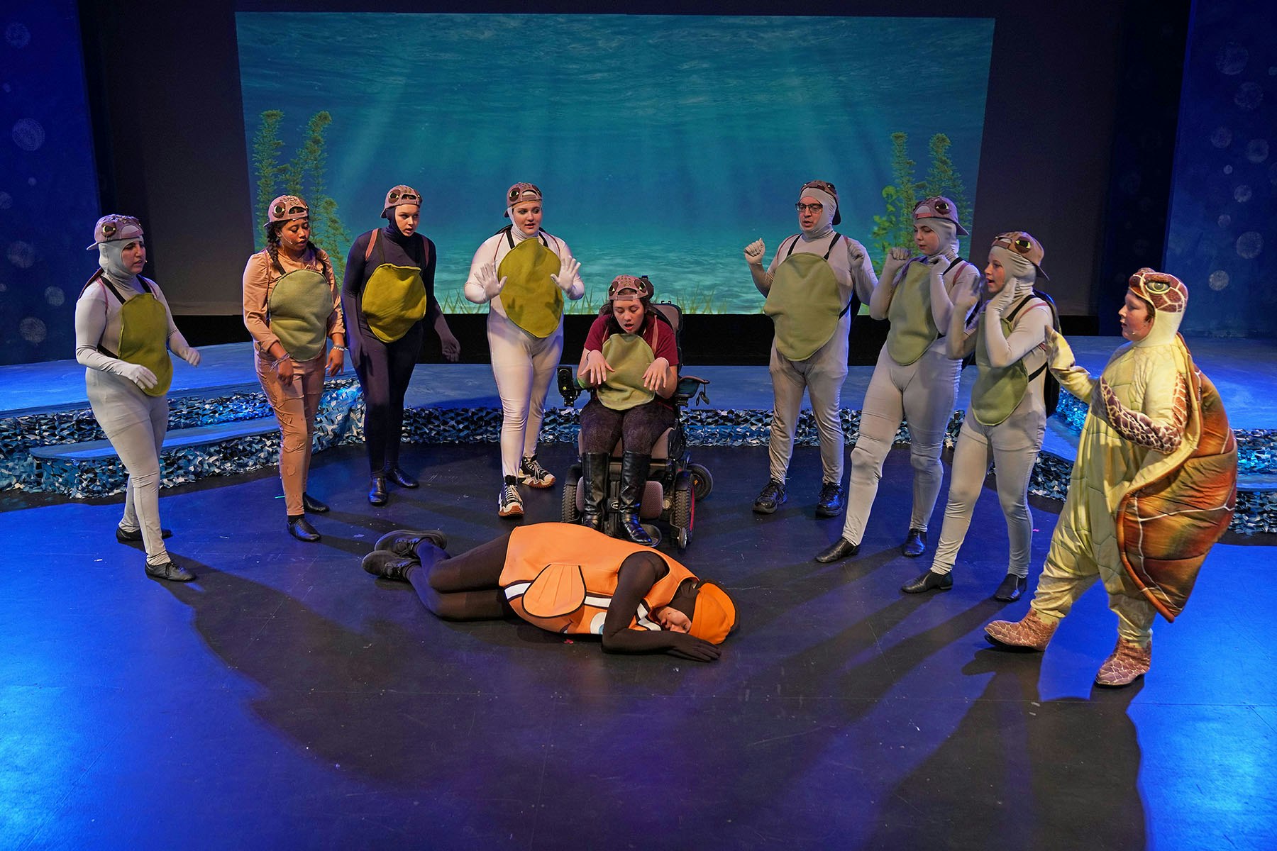 A group of people in costumes resembling sea creatures, with an ocean backdrop, performing a scene.