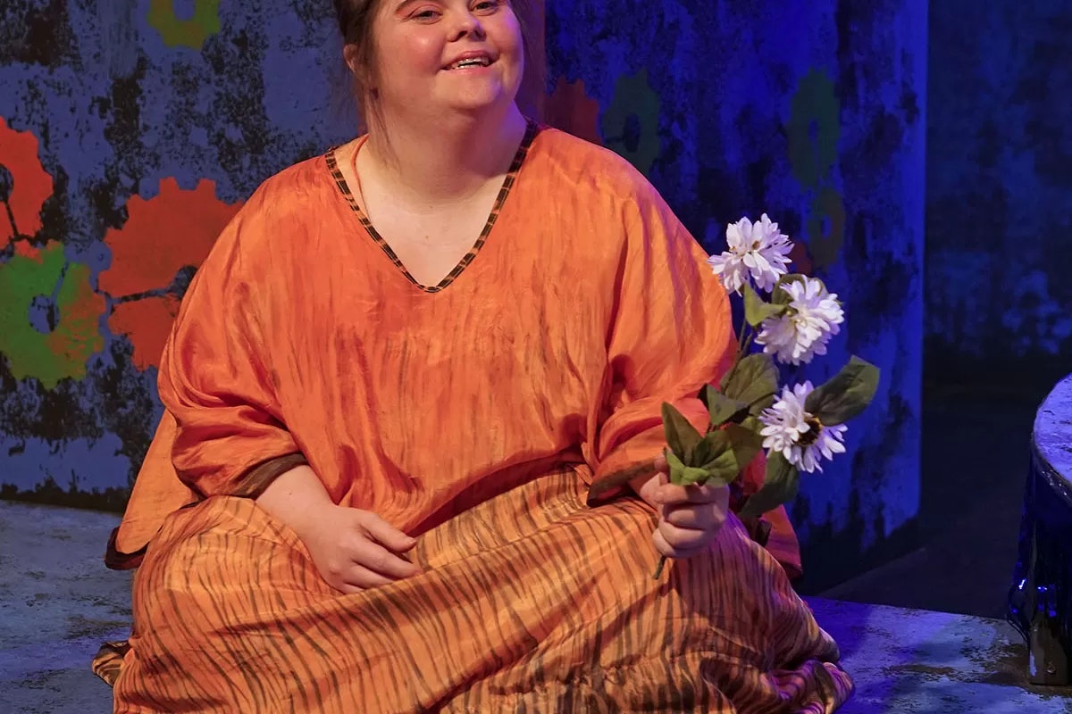 A person in an orange outfit holding white flowers, sitting on a stage with colorful backdrop.
