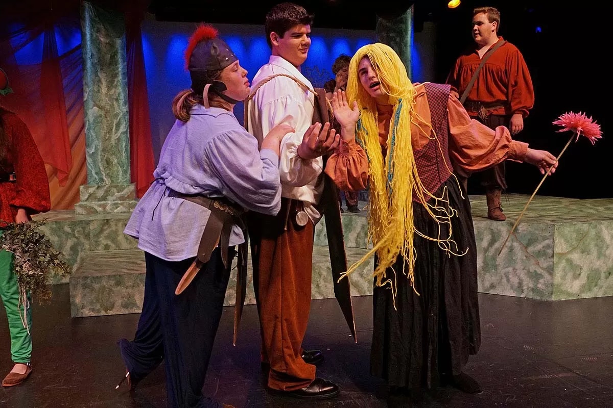 People in costume performing a theatrical play on stage with colorful lighting.