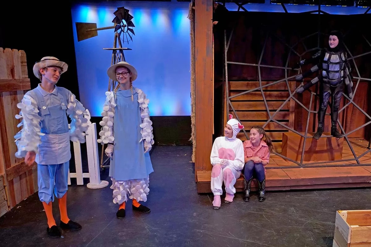 People in a stage play dressed as farm characters, including a spider on a web, a pig, and farmers.