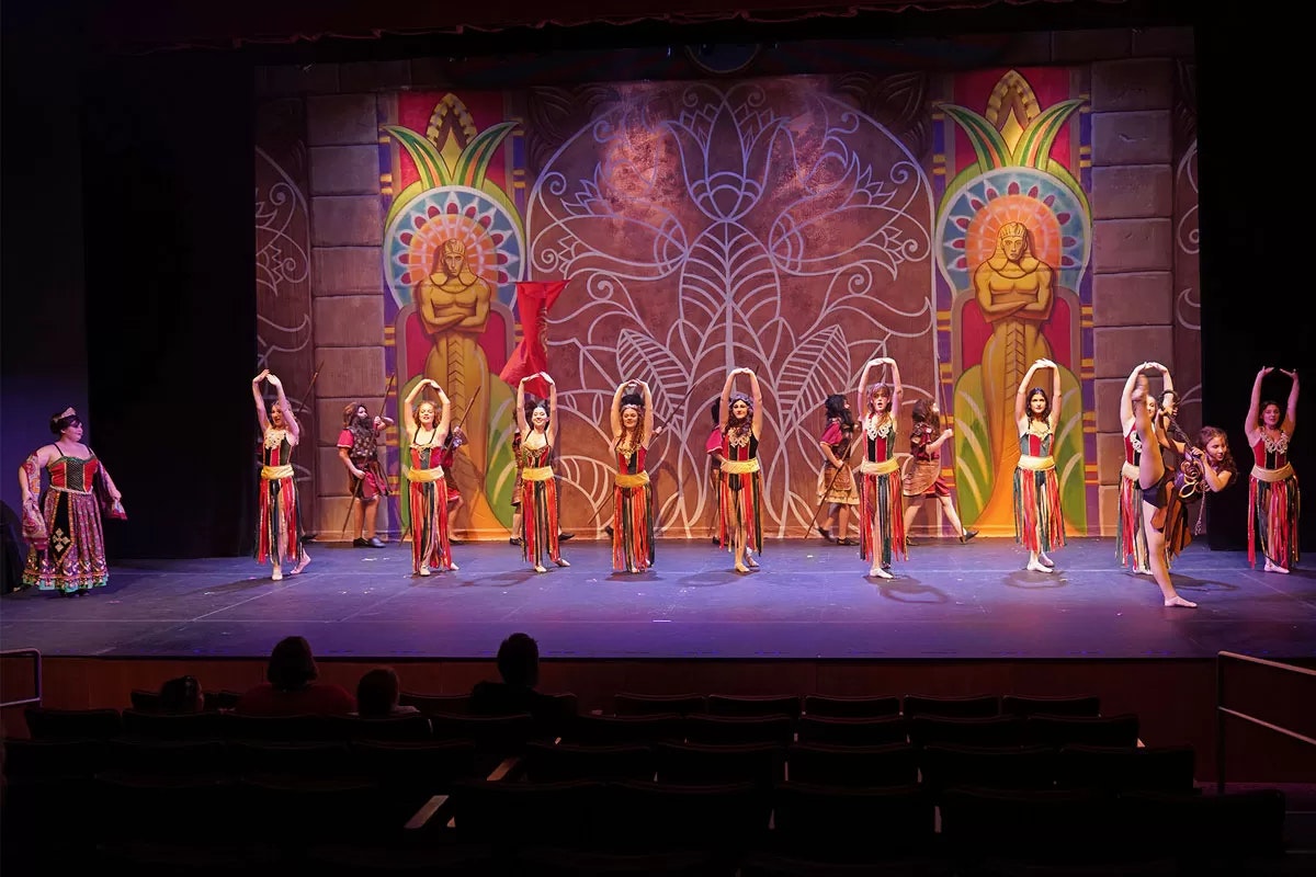 A colorful stage performance with dancers in vibrant costumes, a detailed backdrop, and audience watching.