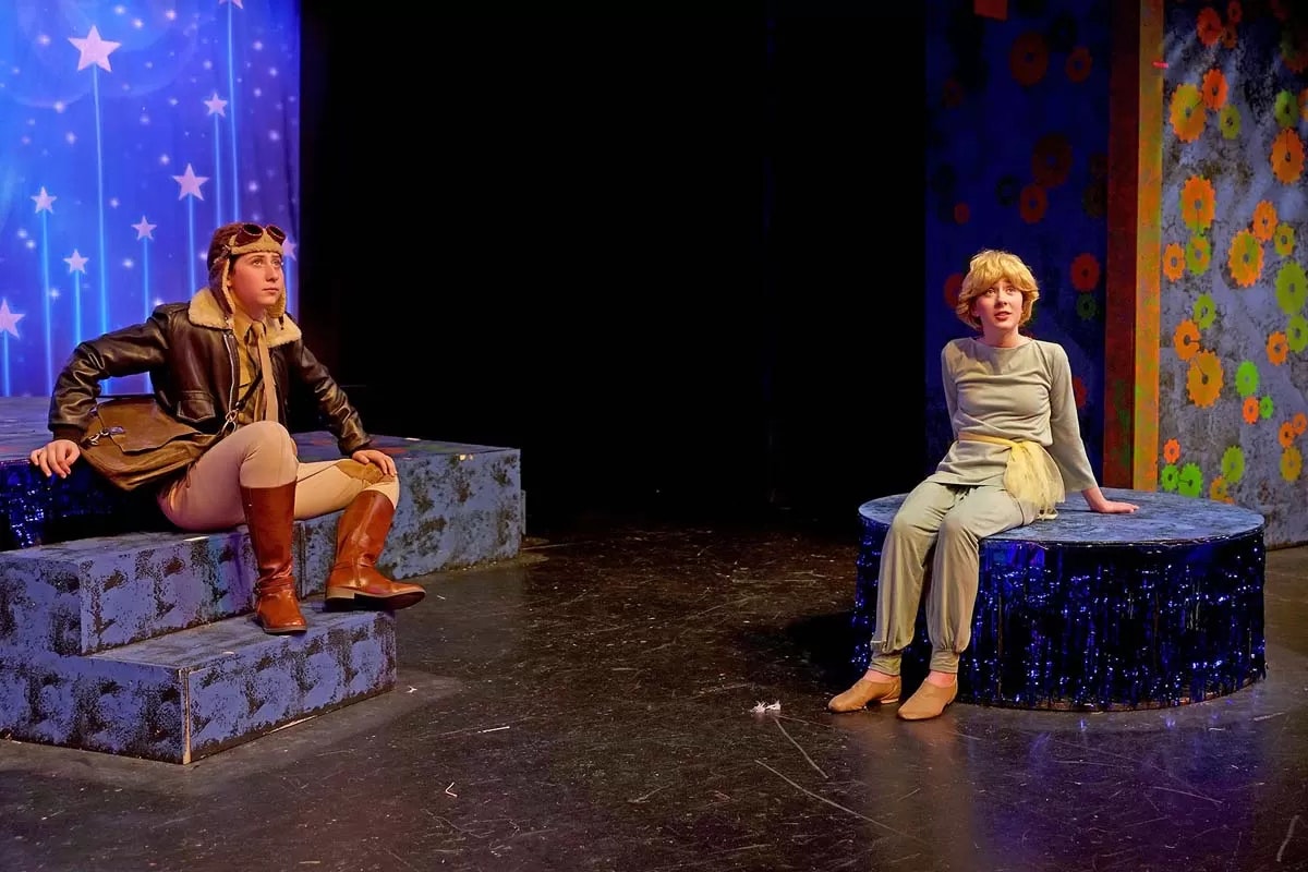 Two actors in a theatrical scene, one in aviator gear and the other in a light-colored costume, with a starry background and floral patterns.