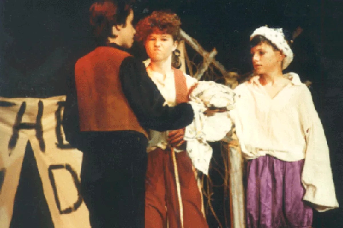 Three people in costume are on stage, likely in a play. One faces away, while the other two are in front of a banner reading "THE...".