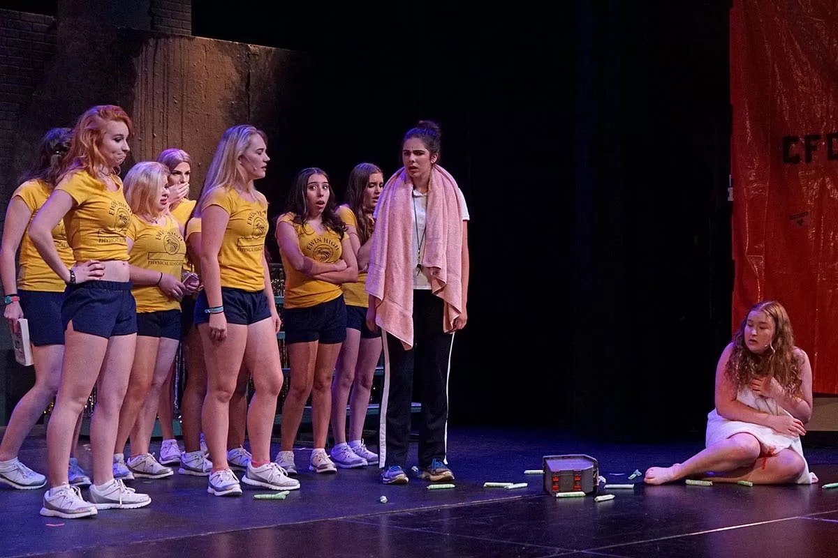 A group of people in yellow shirts stands onstage, looking at a seated person wrapped in a towel.