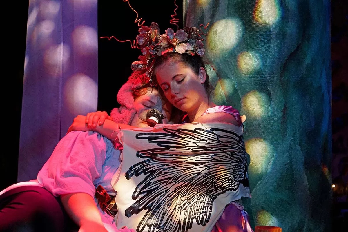 Two people are embracing, one wearing a flower crown and the other a pink costume, with artistic lighting and a serene atmosphere.