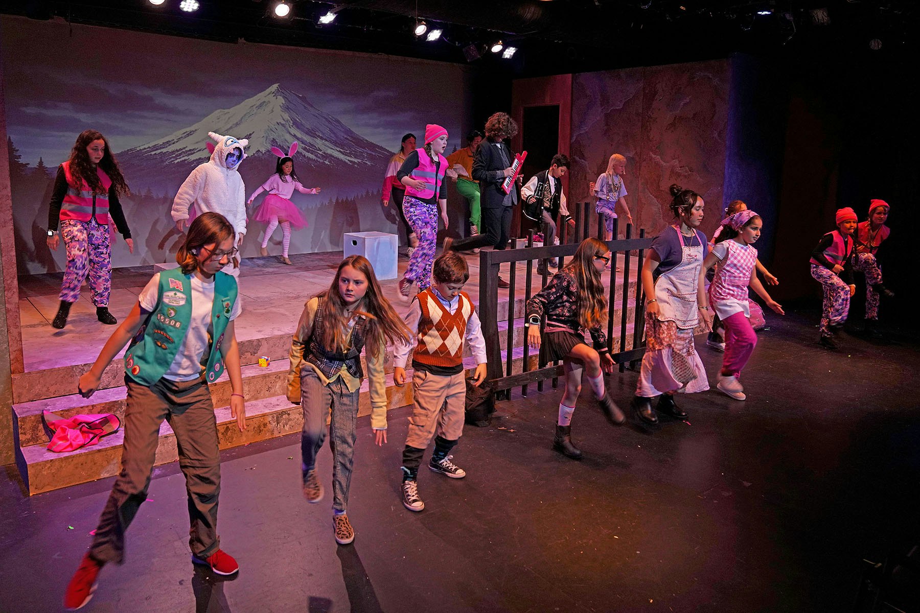 A lively stage performance featuring children in colorful costumes, dancing and acting with a mountain backdrop.