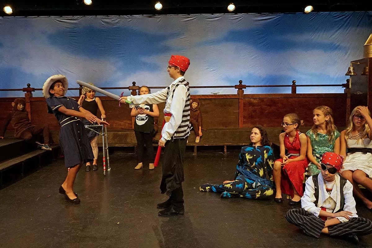 Children in a theater play; a pirate pointing a sword at a person with a hat and others in colorful costumes watching.