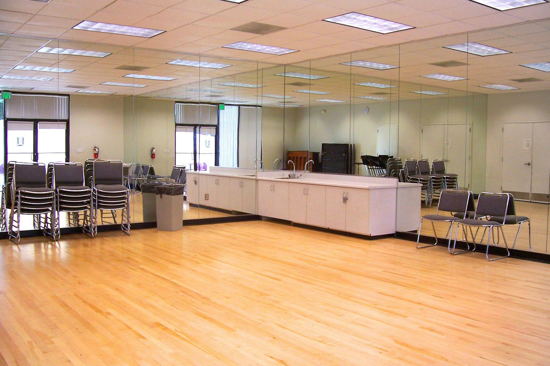 A mirrored room with wooden floors, stacked chairs, a counter with cabinets, a trash bin, and ceiling lights.