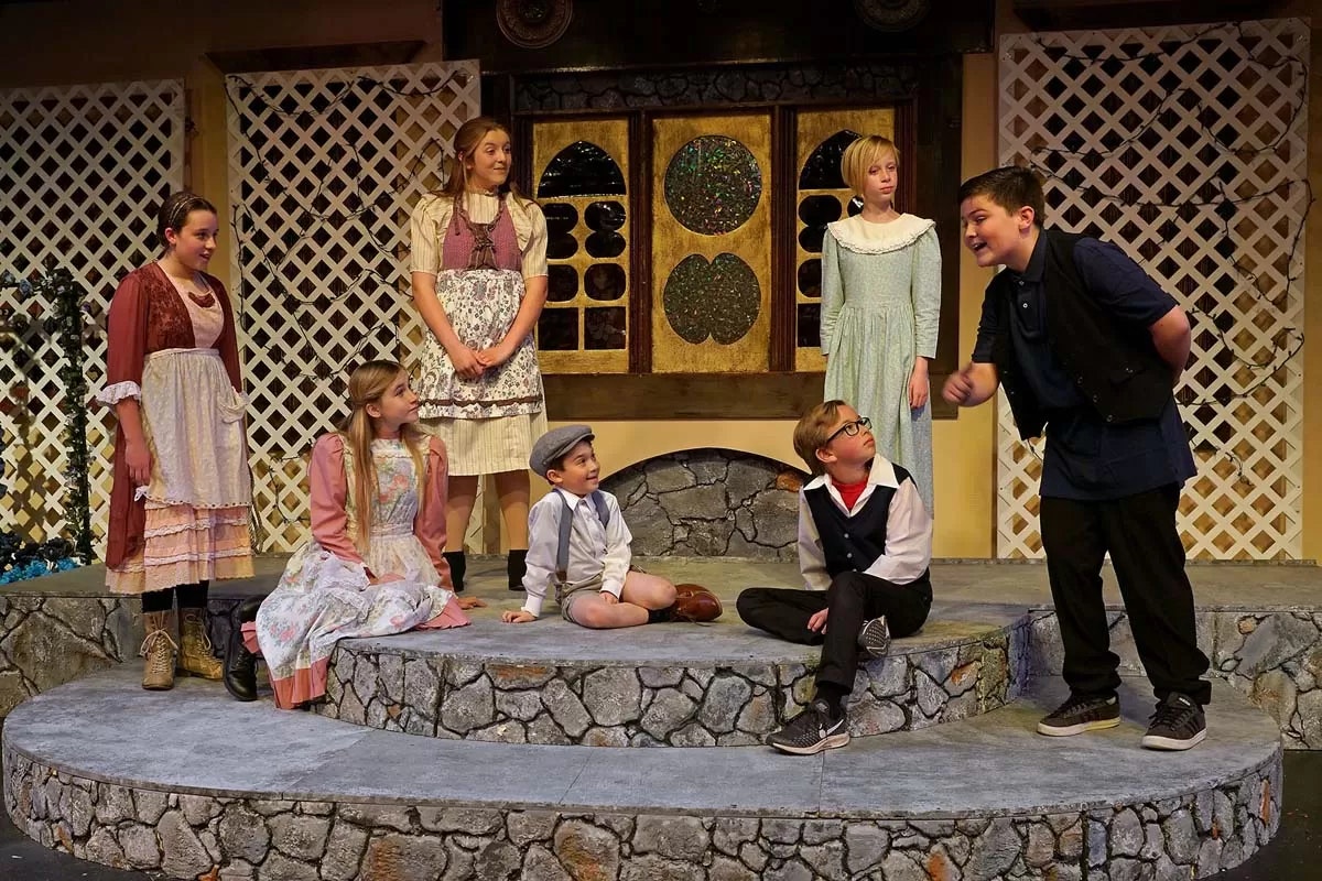 A group of children in period costumes performing a play on a stone stage with a decorative background.