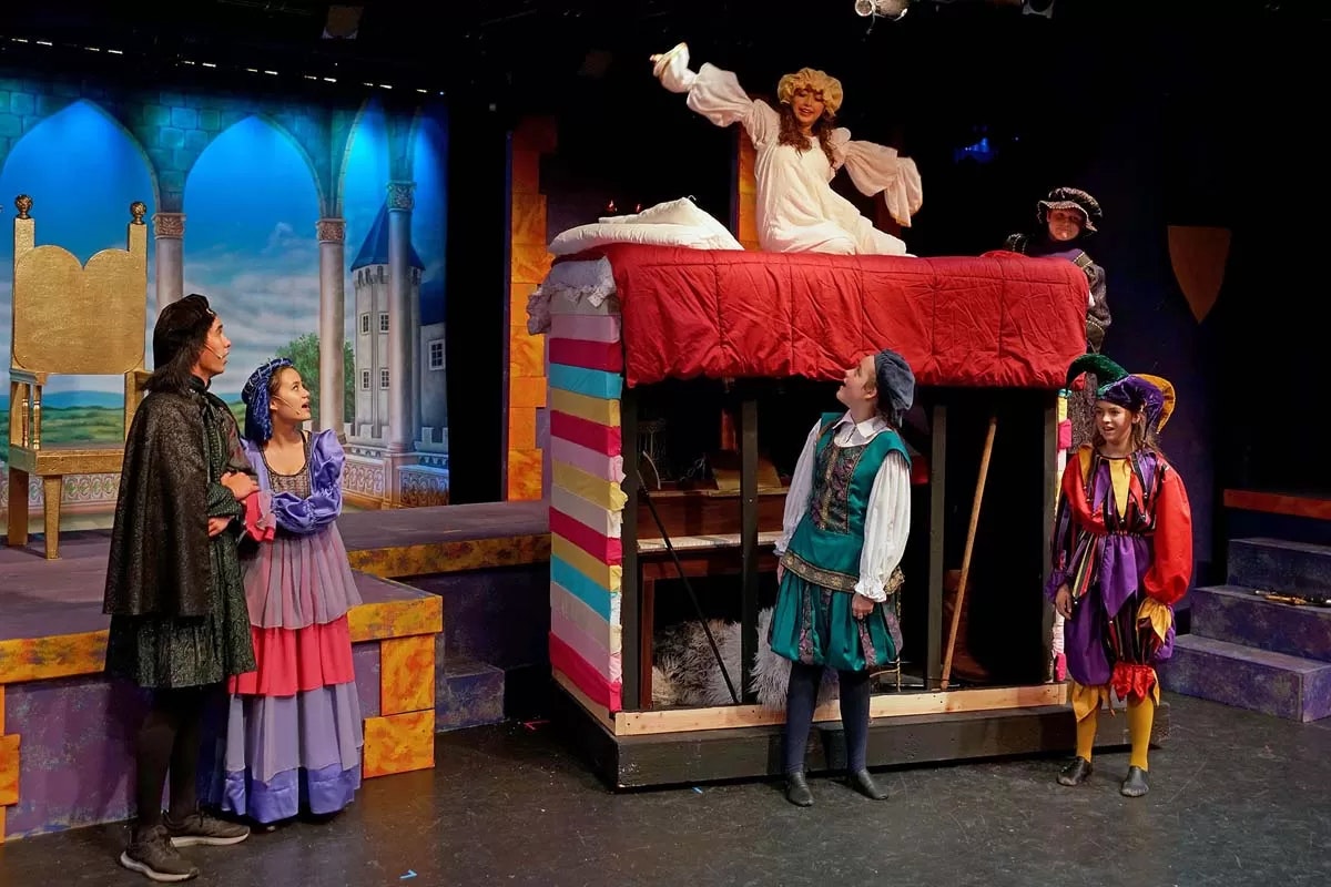 A theater performance scene with colorful costumes, a raised bed, and a backdrop of a castle.