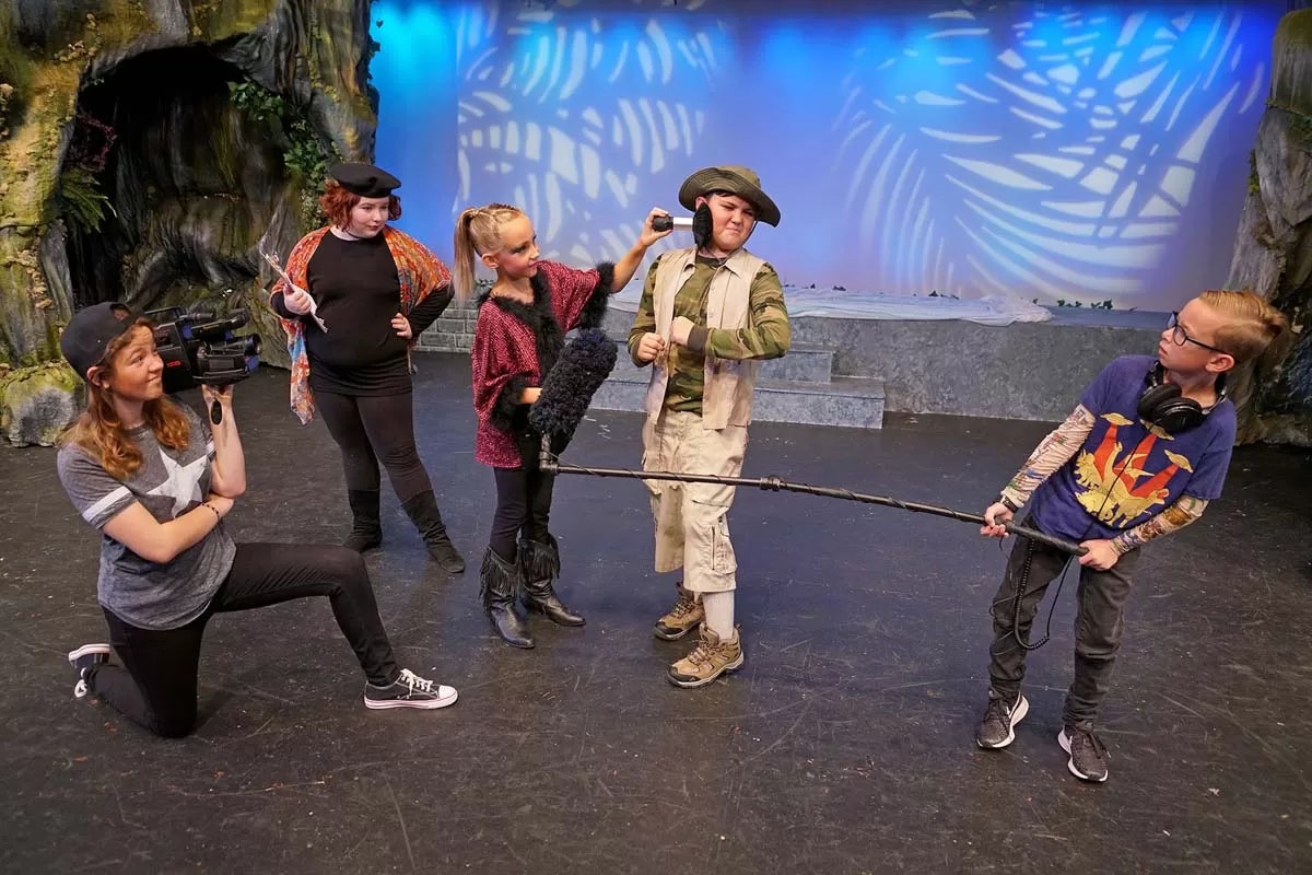 Kids filming a scene on stage with a camera, boom mic, and clapperboard.