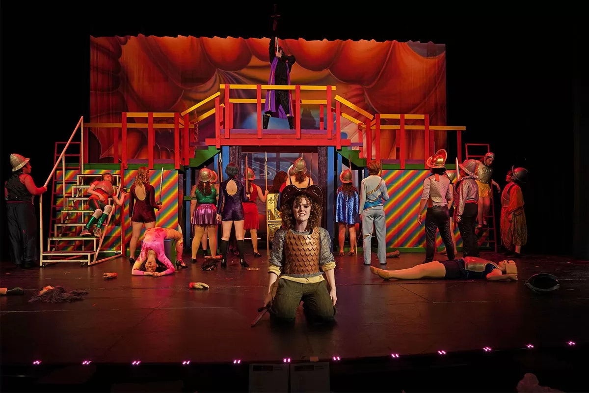A theatrical performance with colorful costumes, a person kneeling center stage, and a vibrant set design featuring a raised platform.
