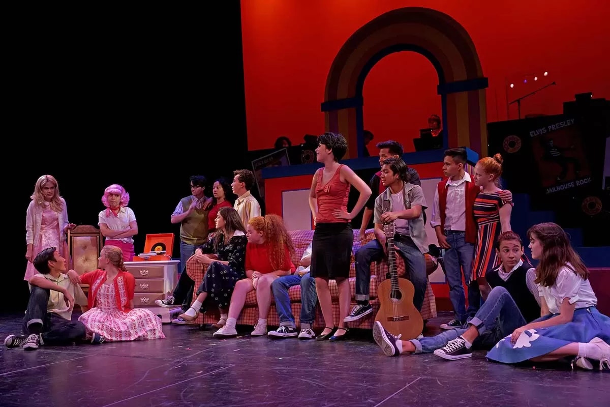 A lively stage performance with actors in retro attire, featuring a diverse group with musical instruments and vibrant backdrop.