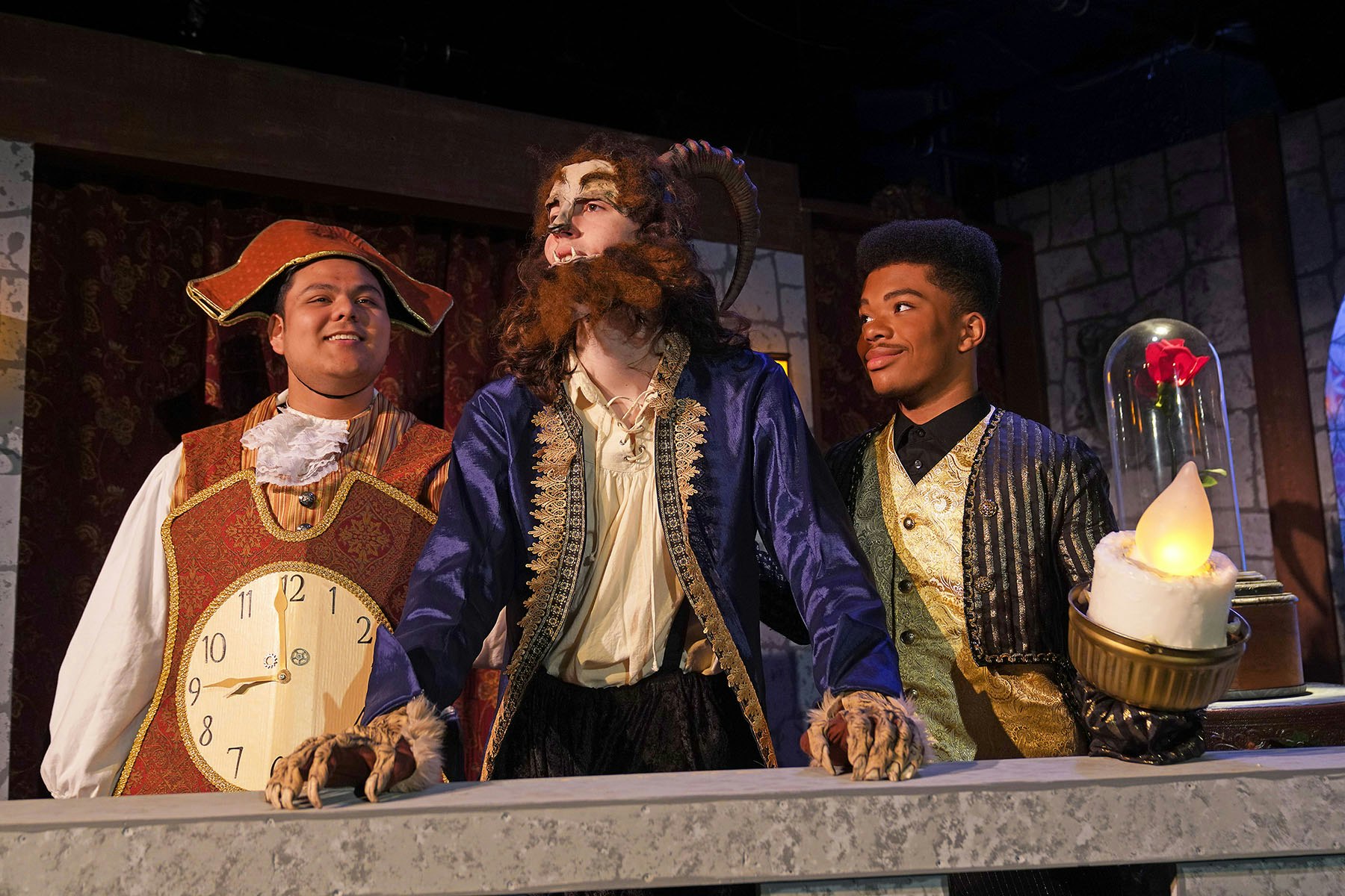 Three people in theatrical costumes: a clock, beast, and a candelabra. A rose under glass is in the background.