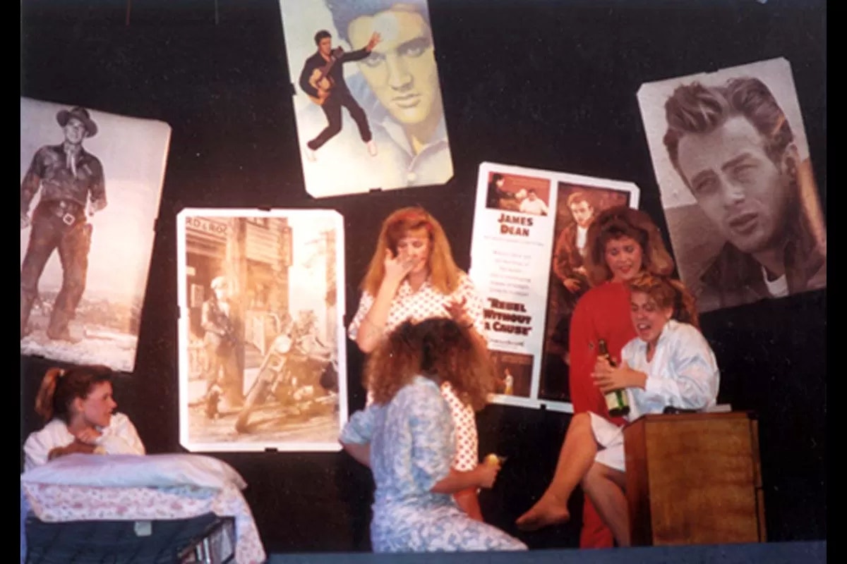 Four people on stage with vintage posters as backdrop, one holding a bottle, others interacting.