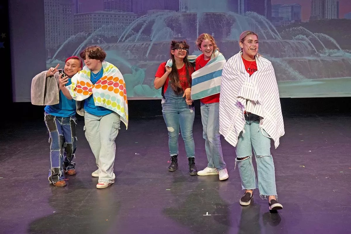 Five people on stage wearing colorful towels, smiling and posing energetically.