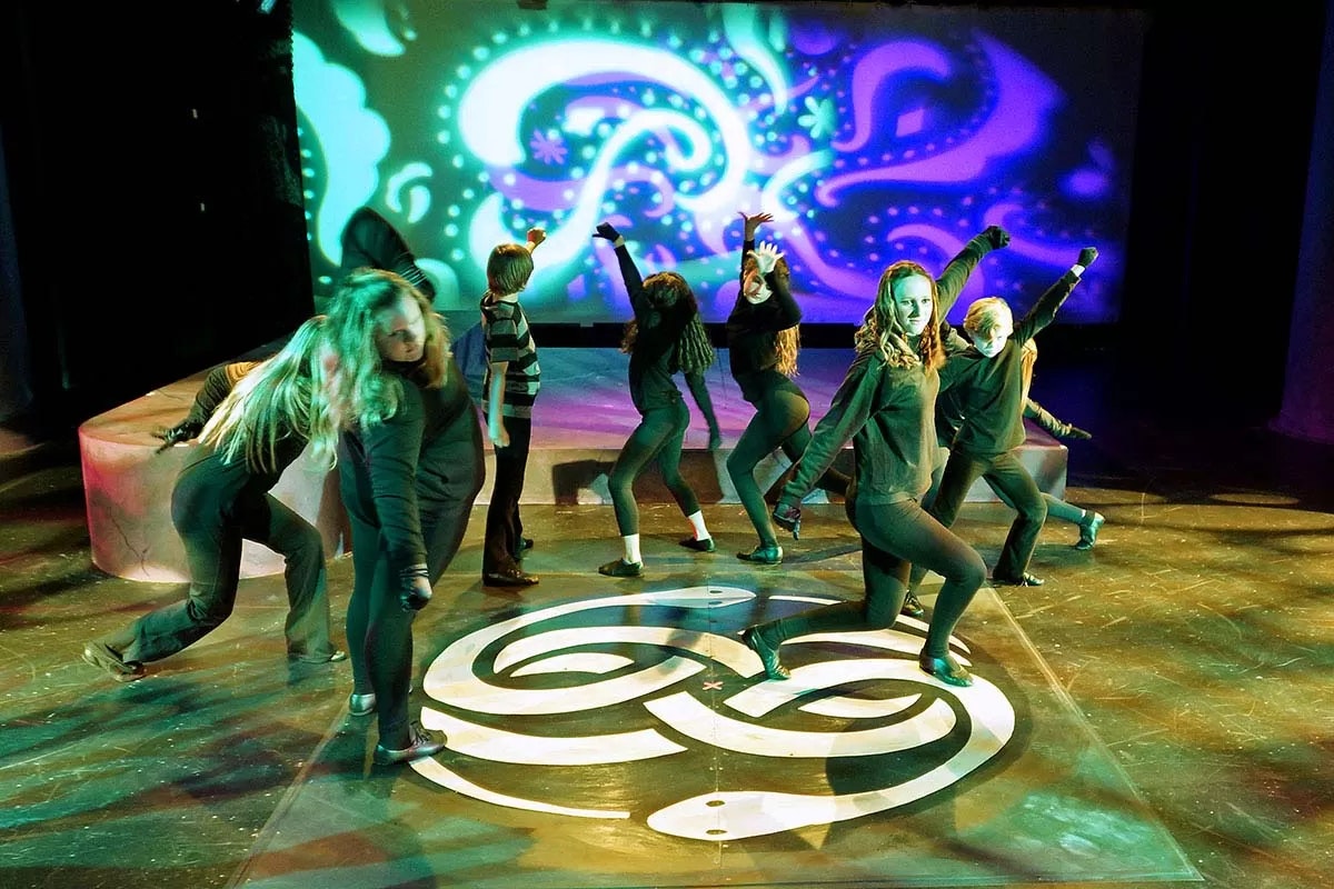 Dancers in black perform energetically on a stage with psychedelic lighting and patterns.
