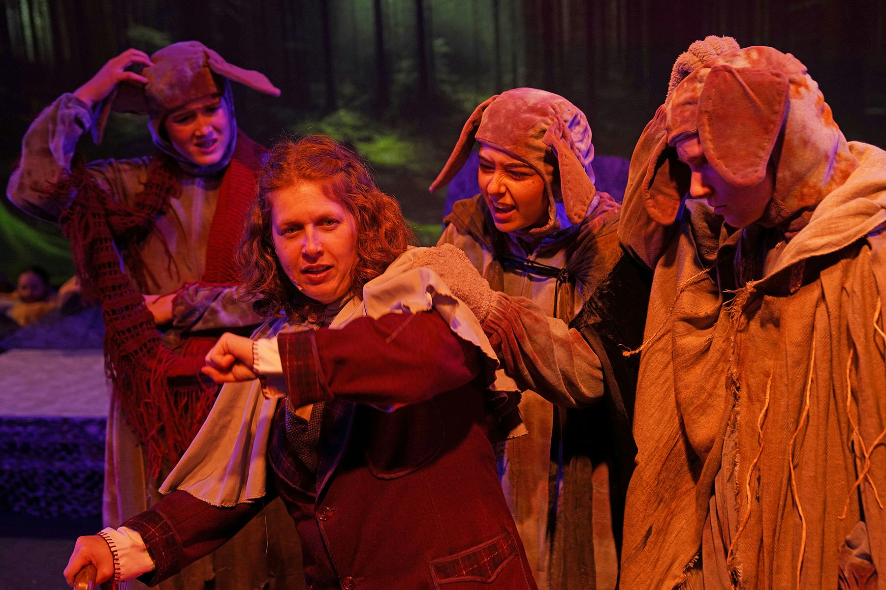 Actors in dog costumes perform a scene on stage in a forest setting with dramatic lighting.