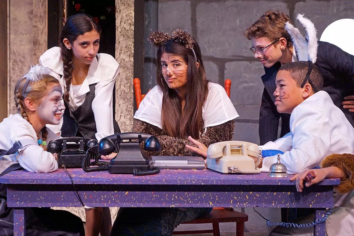 Five people in costume gathered around a table with old telephones, likely performing in a play or theatrical scene.