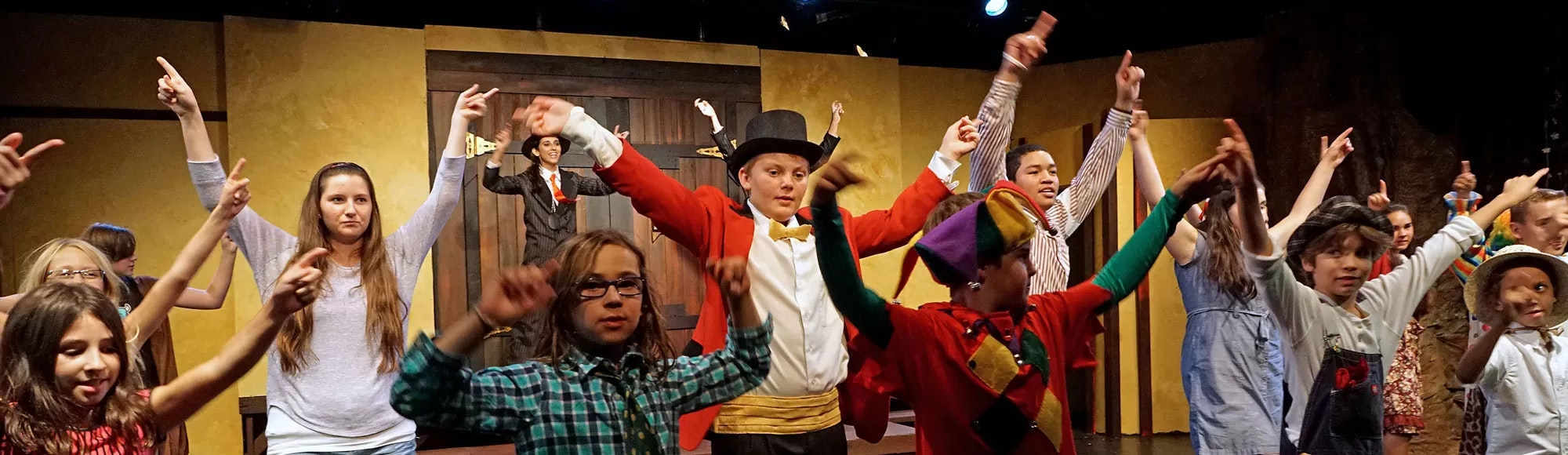 A group of performers, including children, in colorful costumes, raising their arms on stage during a theatrical performance.
