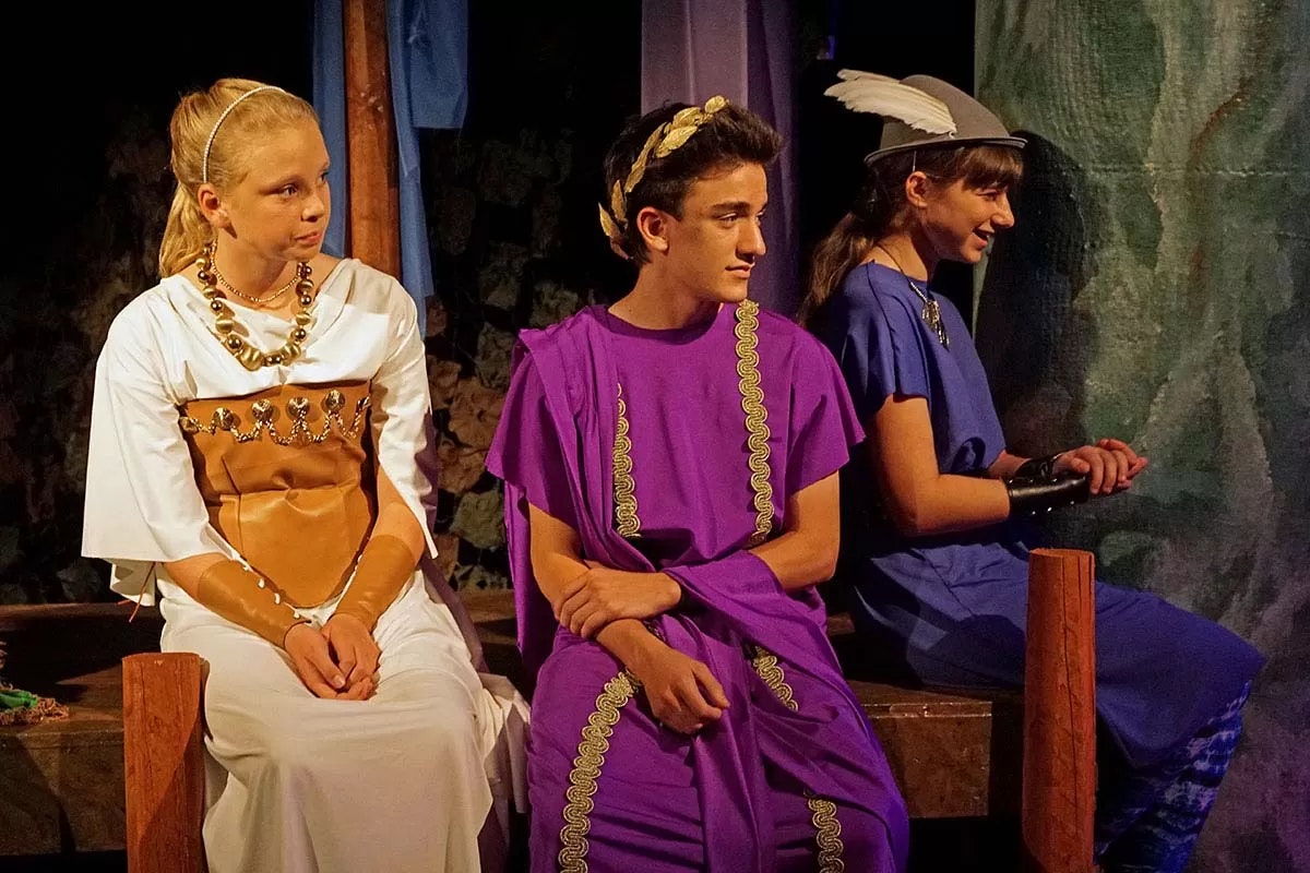 Three people in ancient-style costumes sit on a bench; one in white, one in purple, and one in blue with a feathered hat.