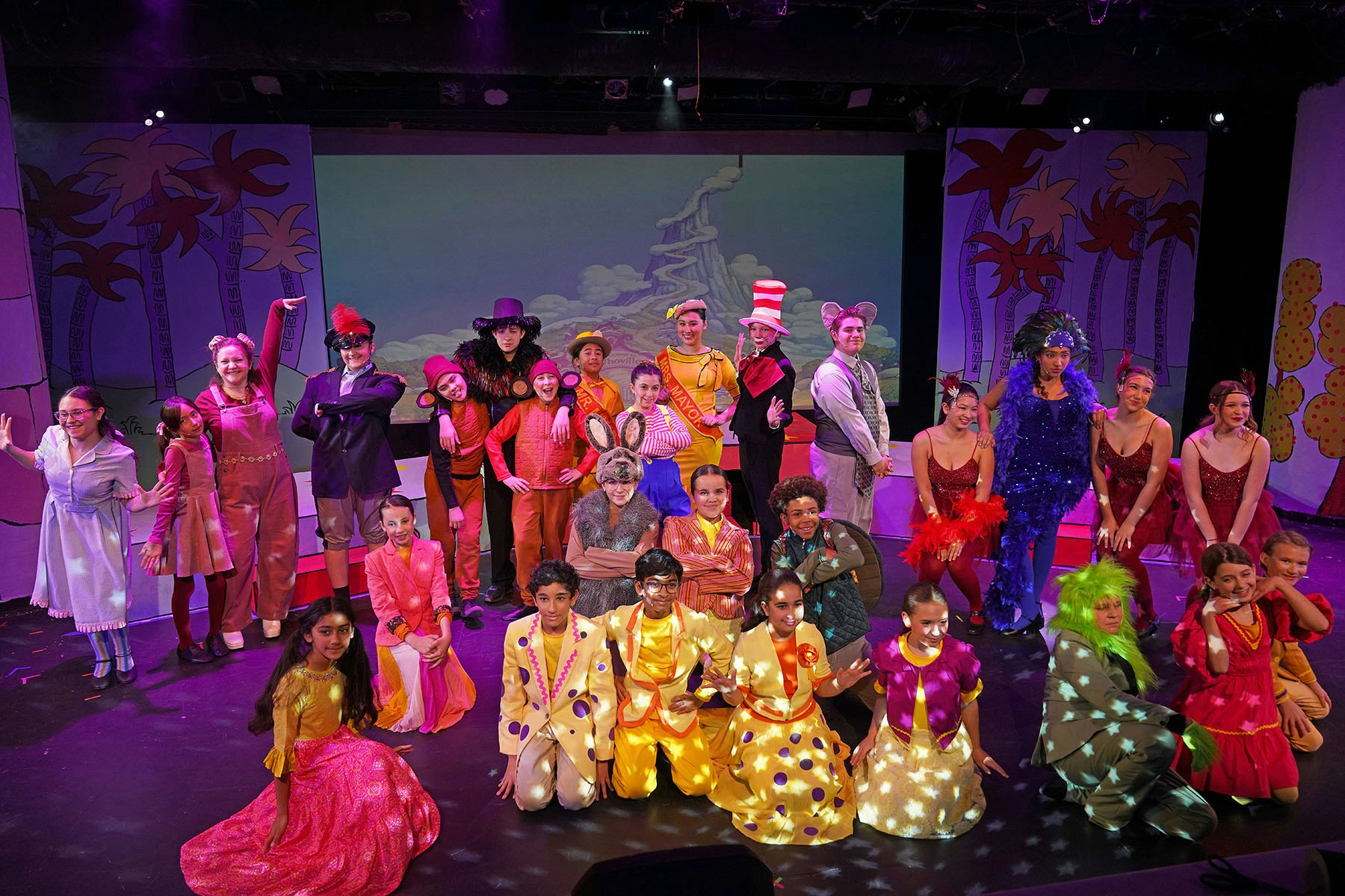 A colorful theatrical performance with a large cast in costume, posing on stage with a vibrant backdrop.