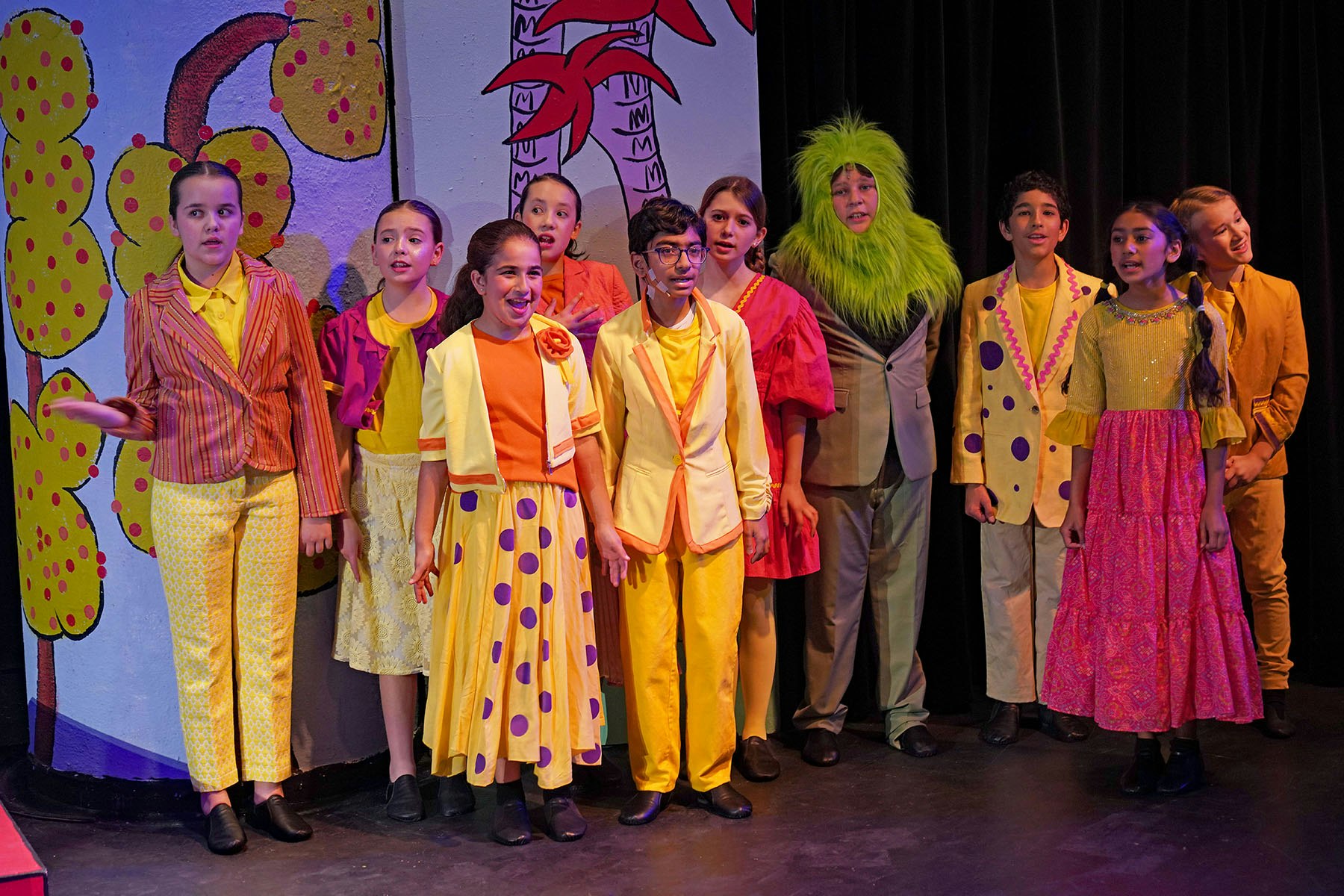 A group of children in colorful costumes perform on stage, with a vibrant backdrop and expressive poses.
