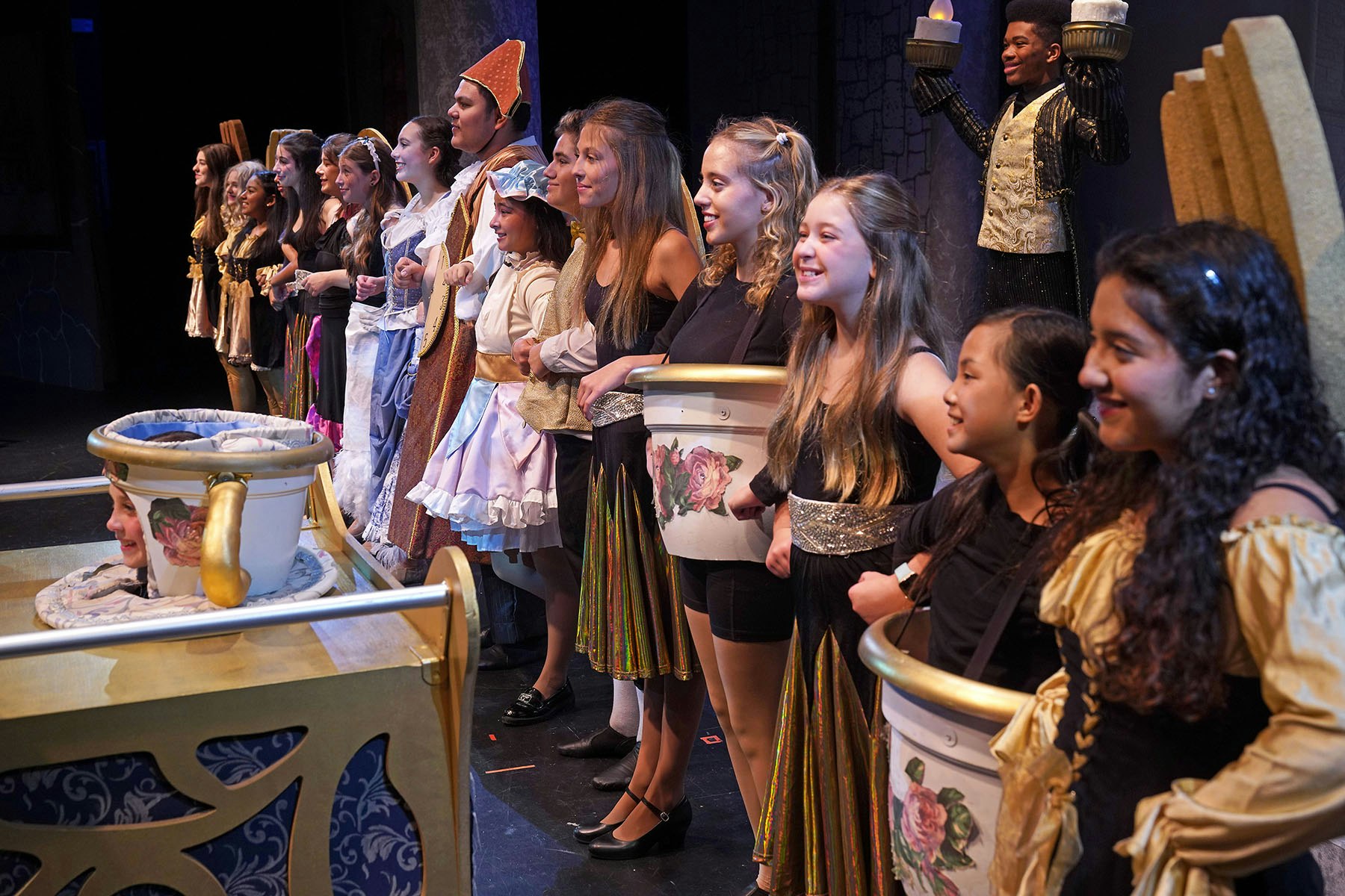 A group of performers in colorful costumes stands on stage, smiling and lined up, likely part of a theatrical production.