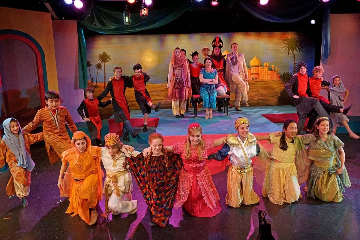 A group of people in colorful costumes performing on stage with a backdrop of a desert scene and palace.