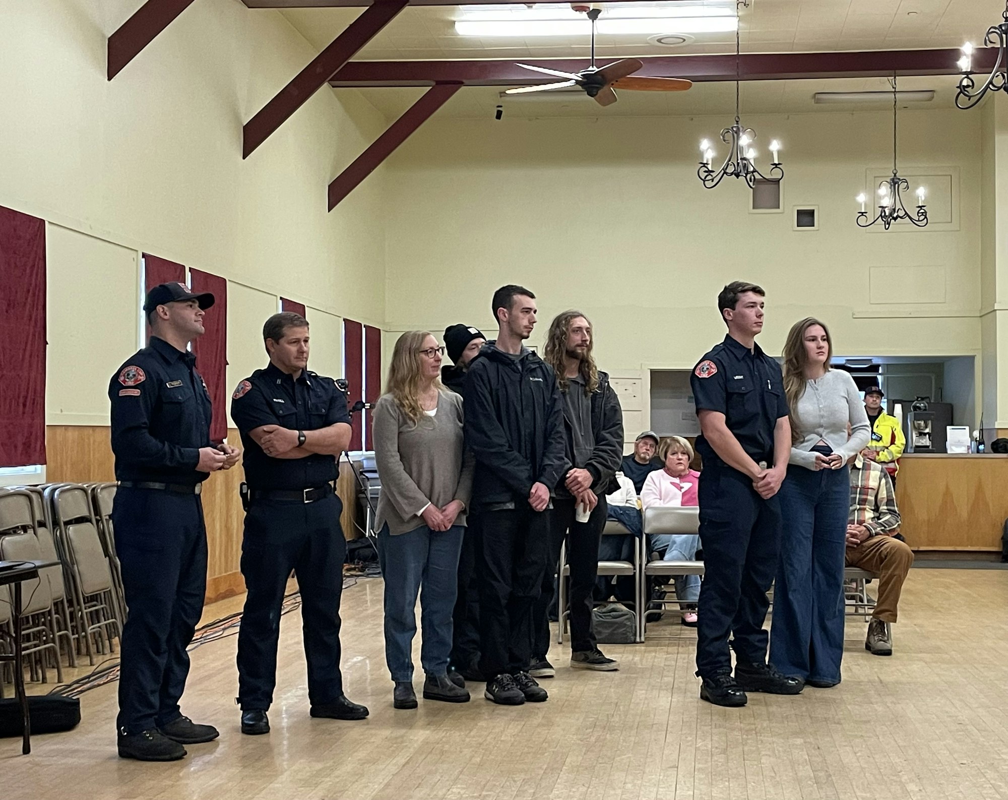 A group of people, including two in uniforms, stand together in a community hall with people seated in the background.