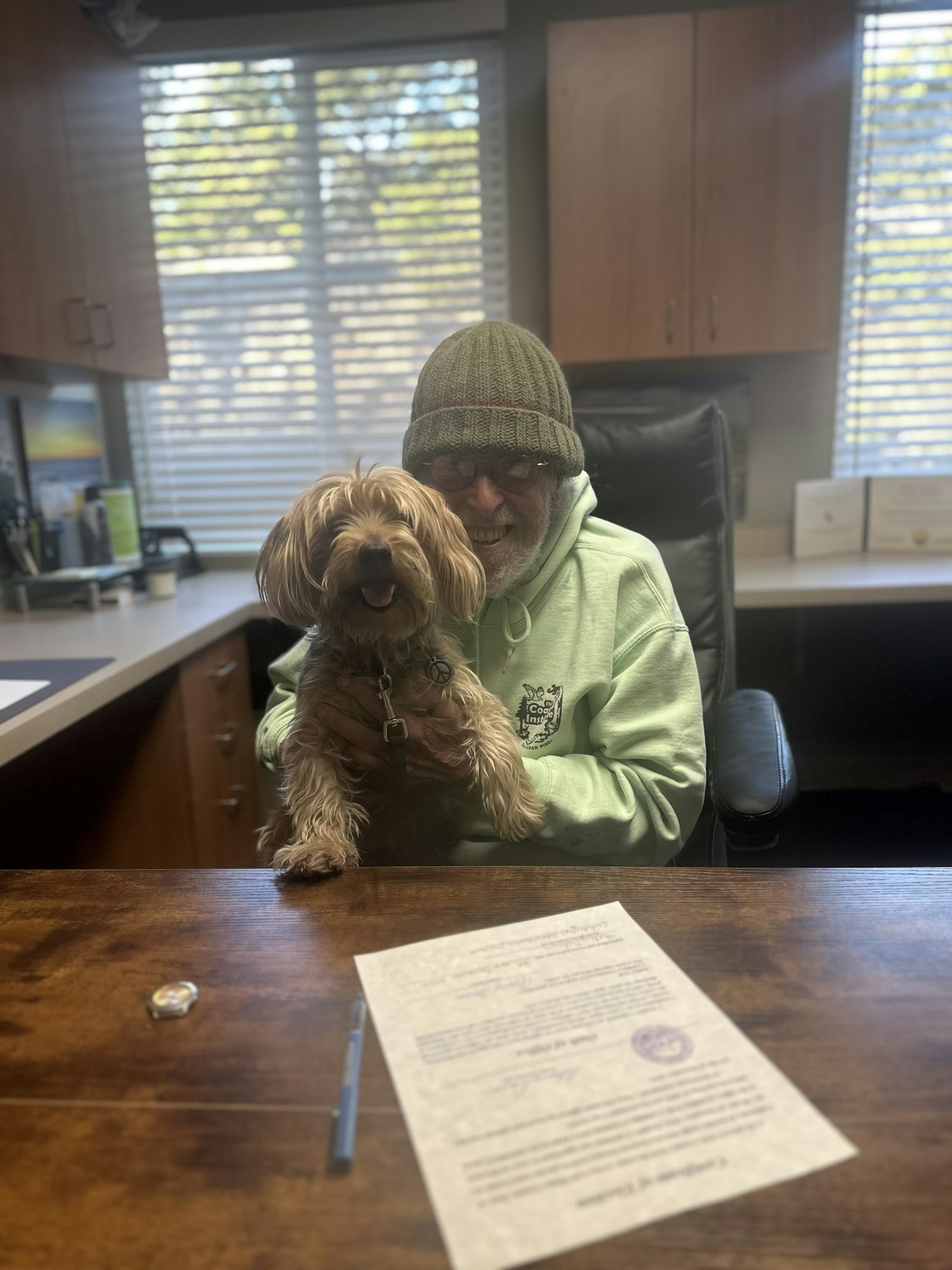 Person in a green hoodie and beanie holding a small, fluffy dog in an office. A document and pen are on the desk.