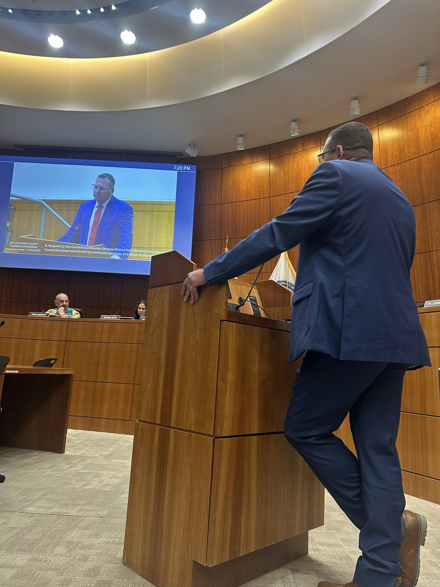 A speaker in a suit appears to be addressing a gathering, with an audience visible in the background.
