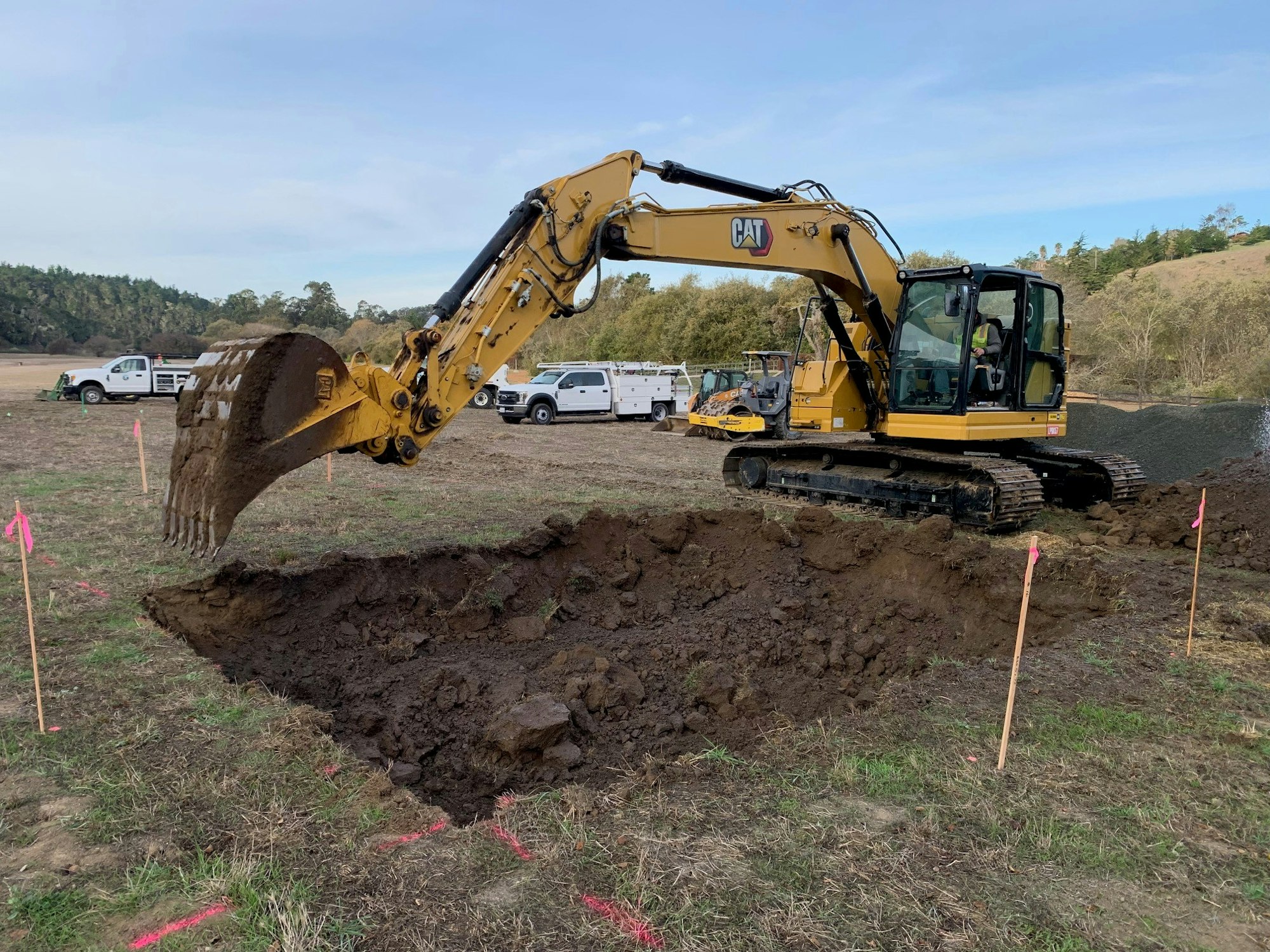 A yellow excavator digging in a field with several trucks in the background.