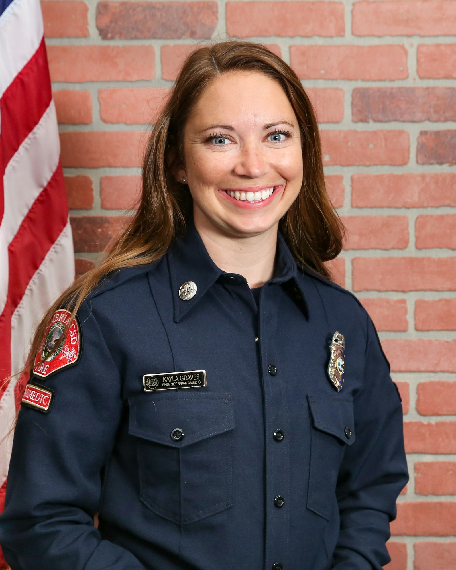 The image features a smiling woman in a navy uniform, standing in front of a brick wall and an American flag.