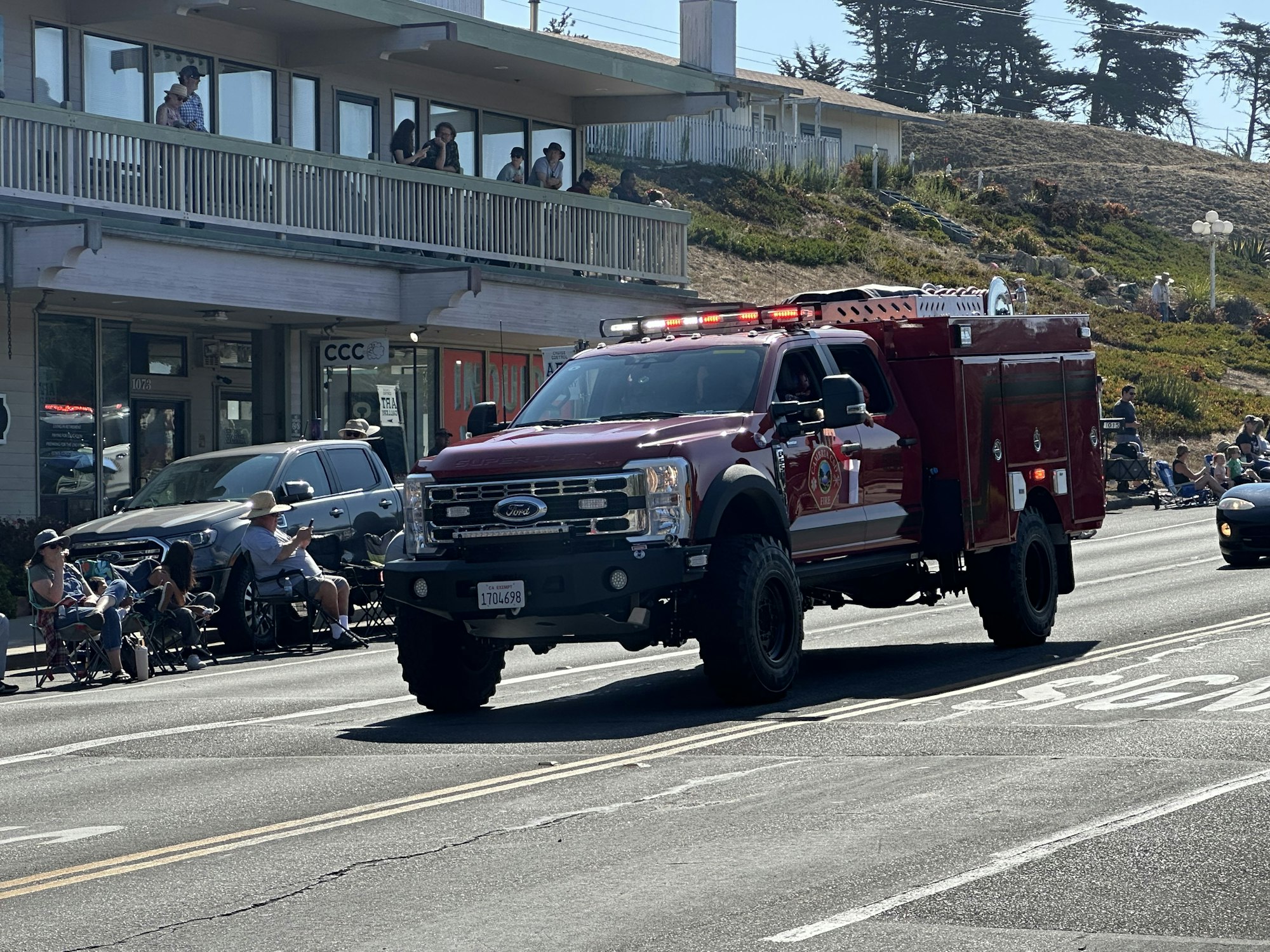 A red fire truck driving on a street, with people watching from the sidewalk and a building in the background.