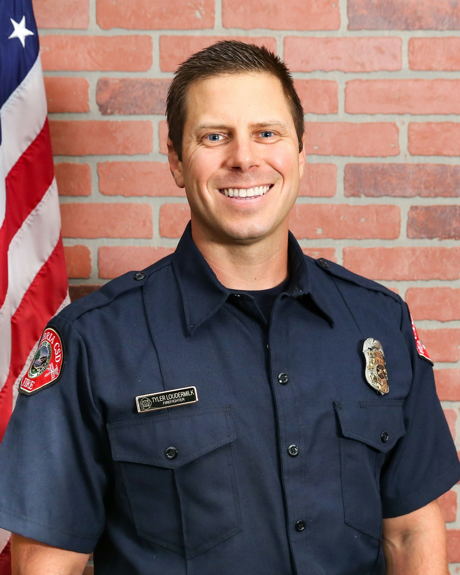 A firefighter in uniform smiling against a brick wall with a flag to the side.