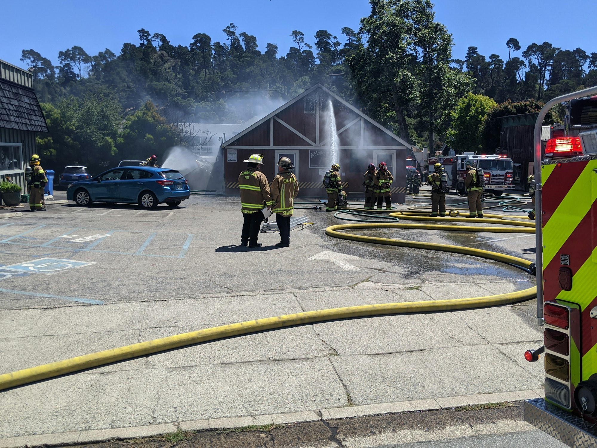Firefighters extinguishing a fire at a building with hoses, smoke visible, surrounded by emergency vehicles and personnel.