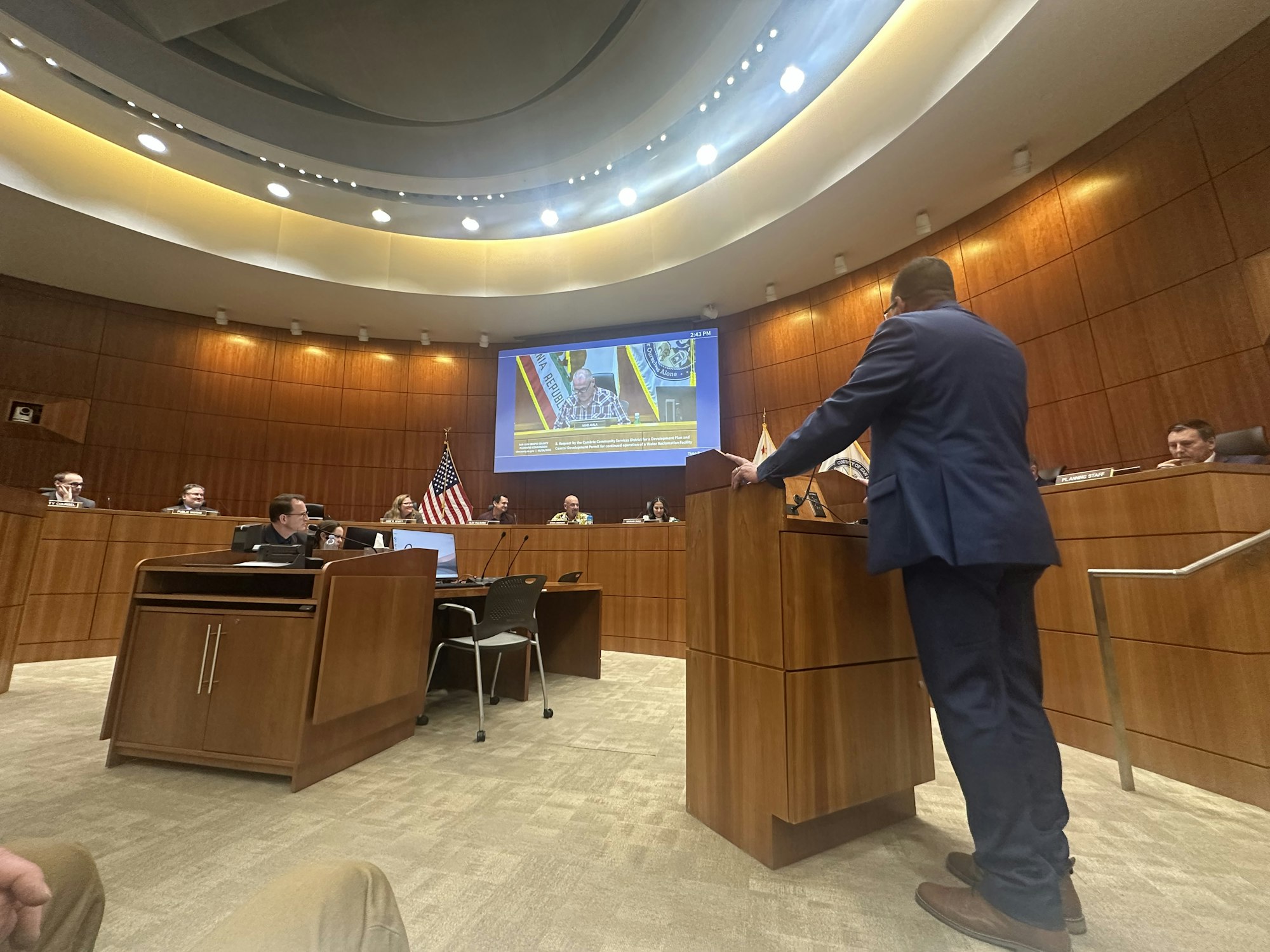 A meeting in a formal chamber, with a speaker at a podium addressing a panel of officials, displaying screens in the background.