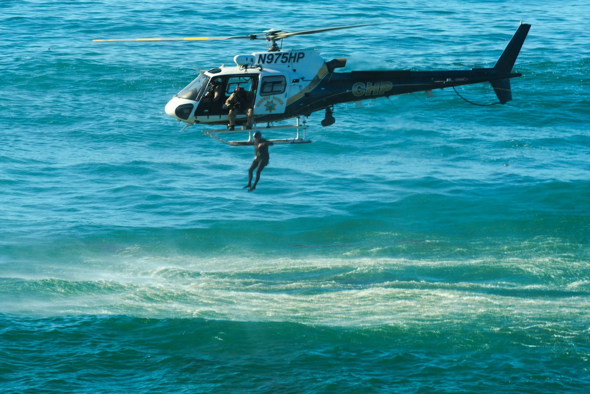A helicopter hovers over the ocean, lowering a rescuer towards the water, likely for a rescue operation.