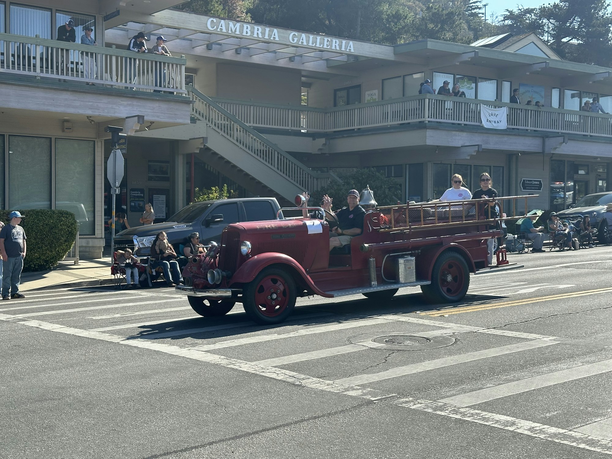 Red vintage fire truck in a parade with people watching from the street and building balcony.