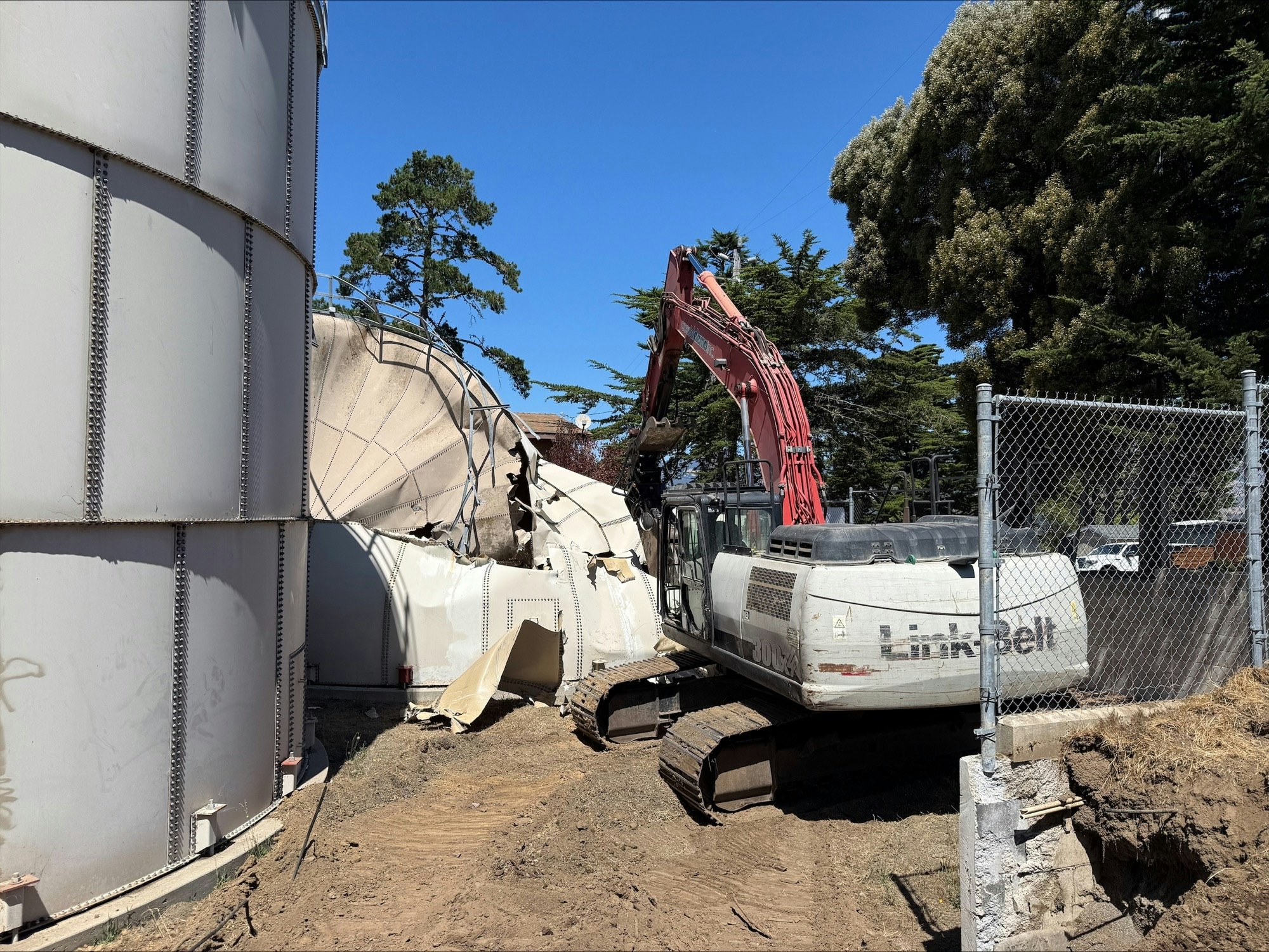 An excavator is demolishing a large metal tank next to trees and a fenced area.