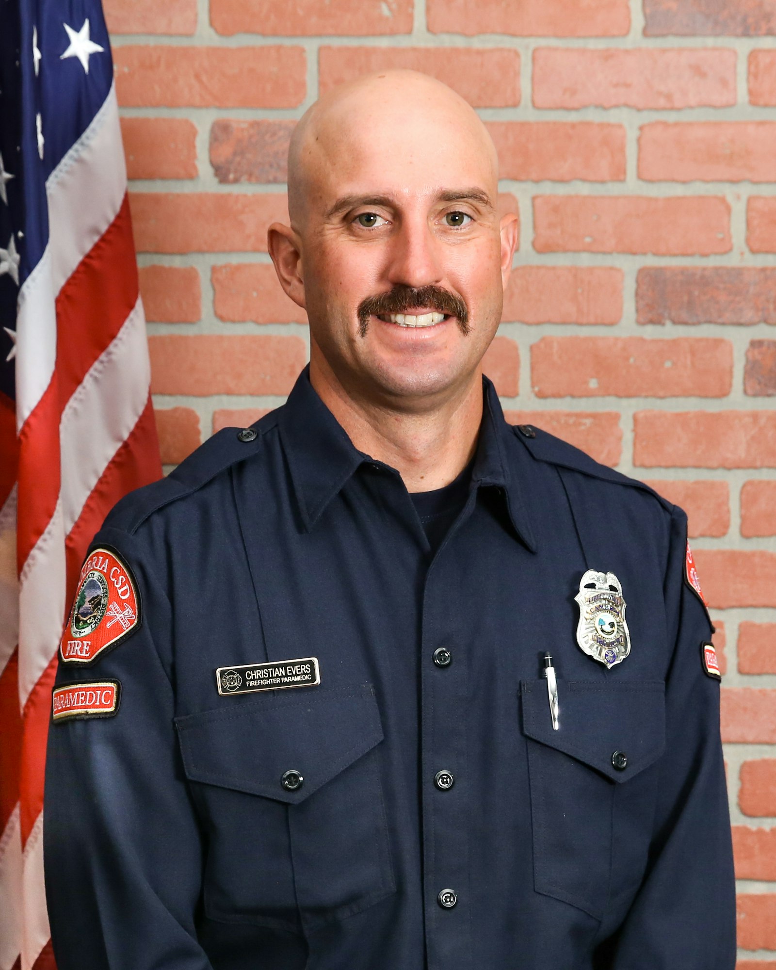 A smiling paramedic poses in uniform in front of a brick wall, with an American flag in the background.